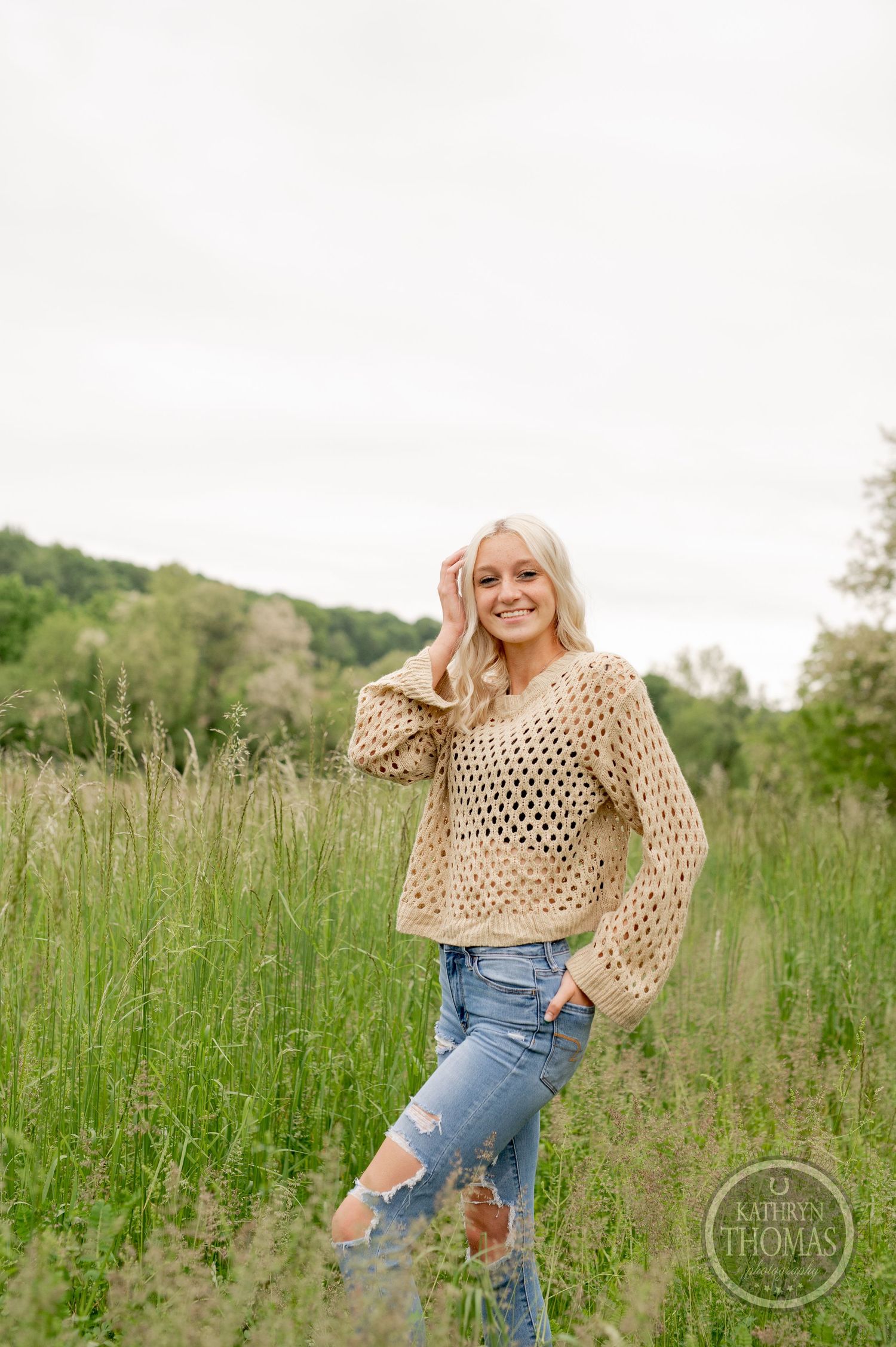 Casual summer outfit with beige crochet sweater and distressed jeans in a sunlit meadow with rolling hills.
