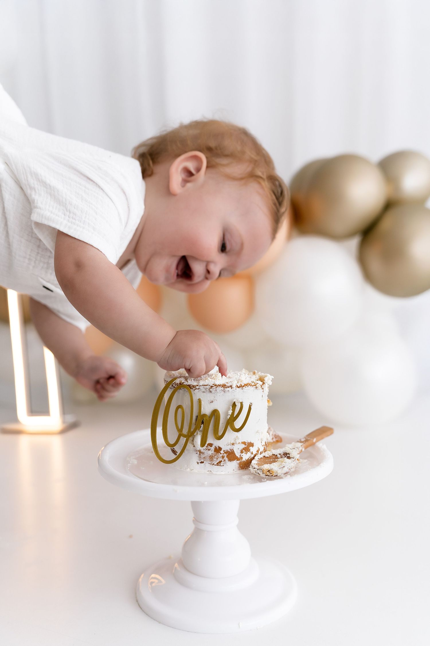 Baby reaching for small white cake with gold 'one' decoration on cake stand.