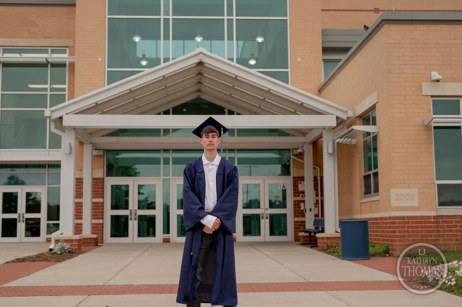 Graduate in cap and gown standing in front of modern school building entrance.