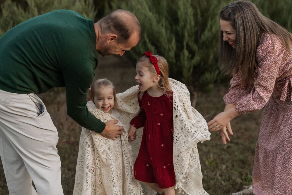 A family shares a playful moment together outdoors in coordinated white, red and pink outfits against an evergreen backdrop.