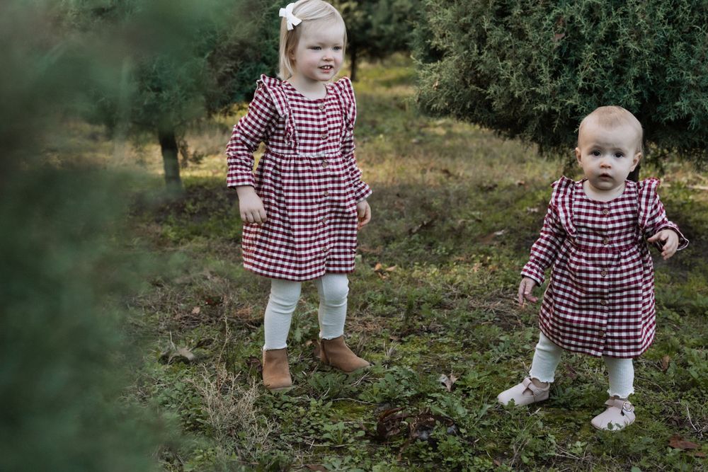 Two small children wearing matching red and white gingham dresses and leggings play together outdoors in nature.