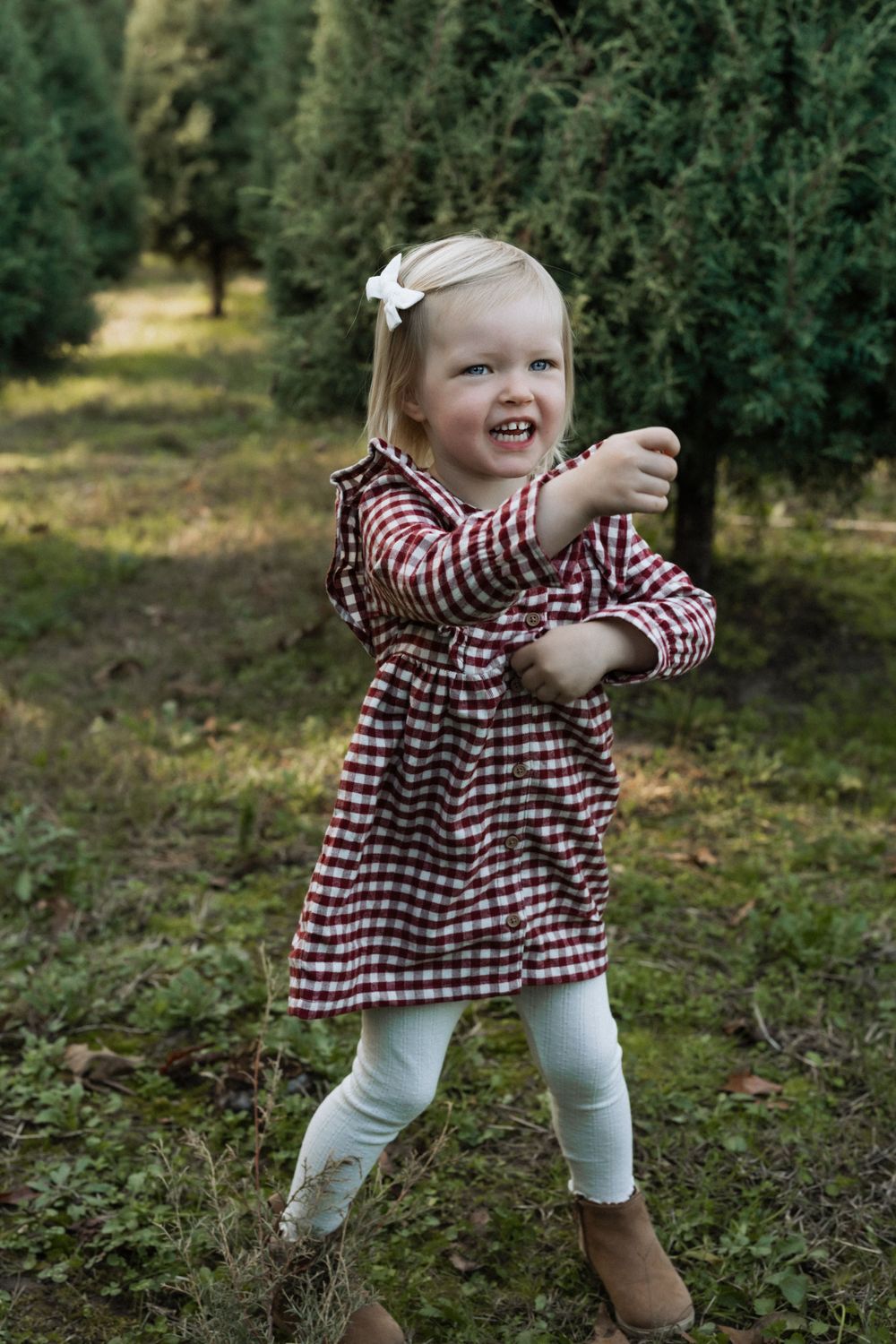 A child in a red gingham dress and white leggings laughs while running through an evergreen tree farm.