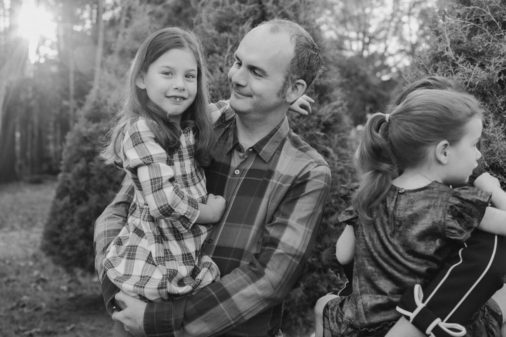 A black and white family portrait taken outdoors in a woodsy setting with sun flare coming through the trees.