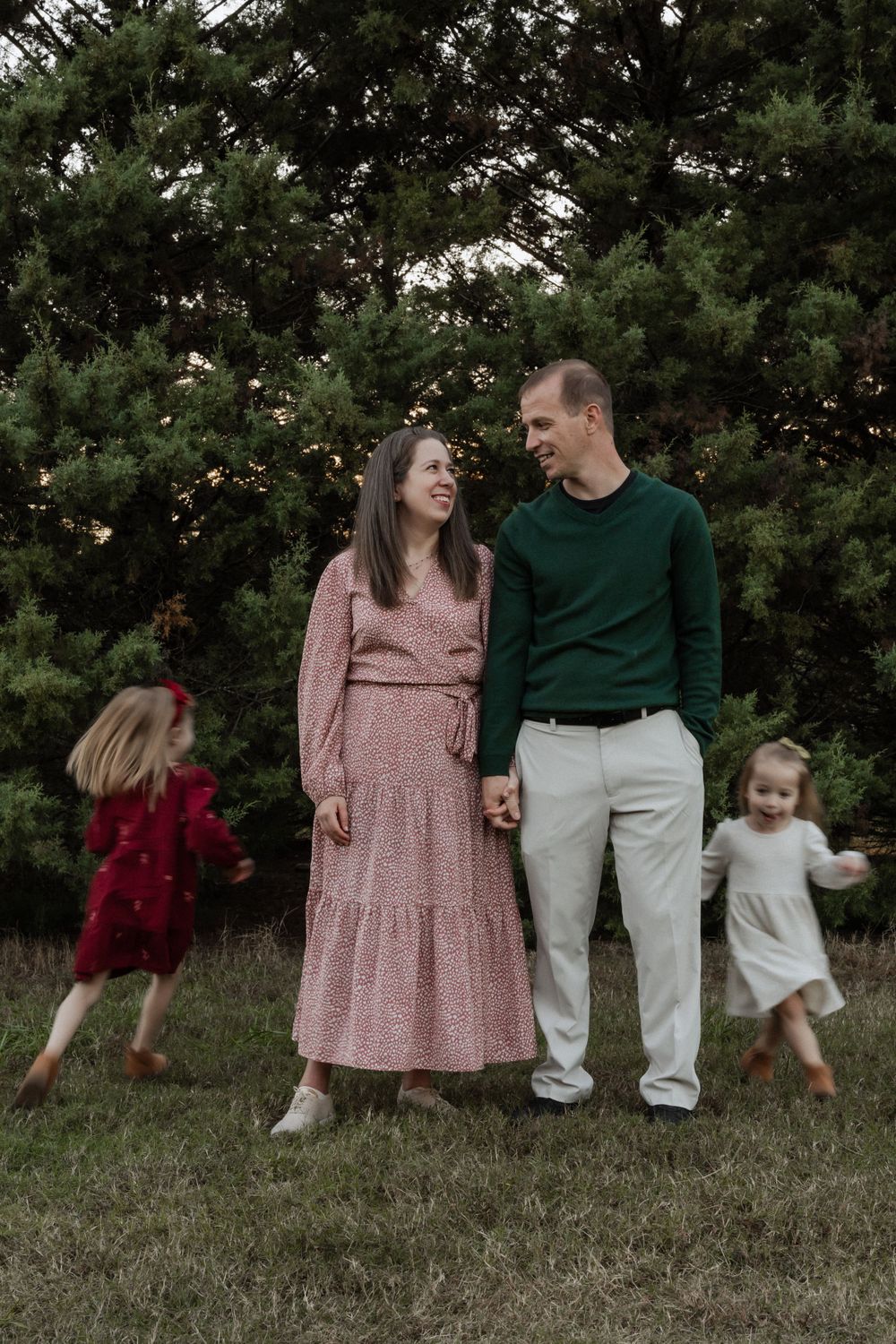 A joyful family moment captured outdoors with two playful children running while parents share a laugh by evergreen trees.