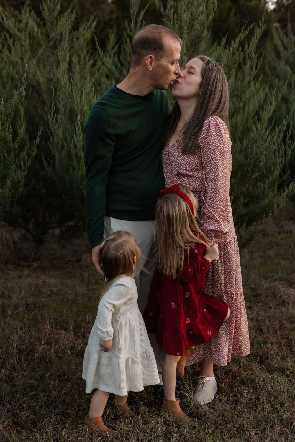 A couple shares a tender moment while two young children in dresses stand beside them against an evergreen backdrop.