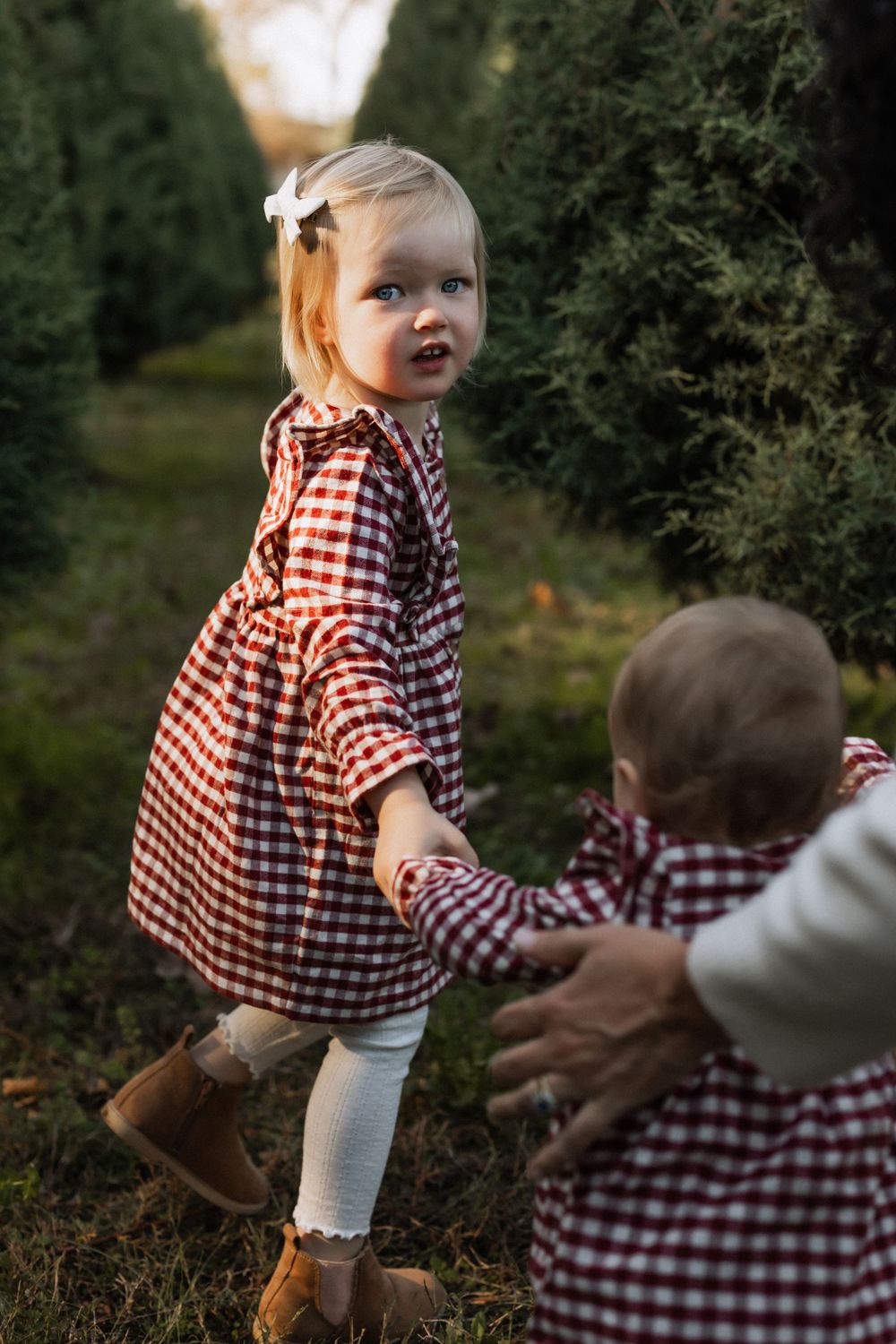 Children wearing matching red and white gingham outfits play together among green evergreen shrubs in an outdoor setting.