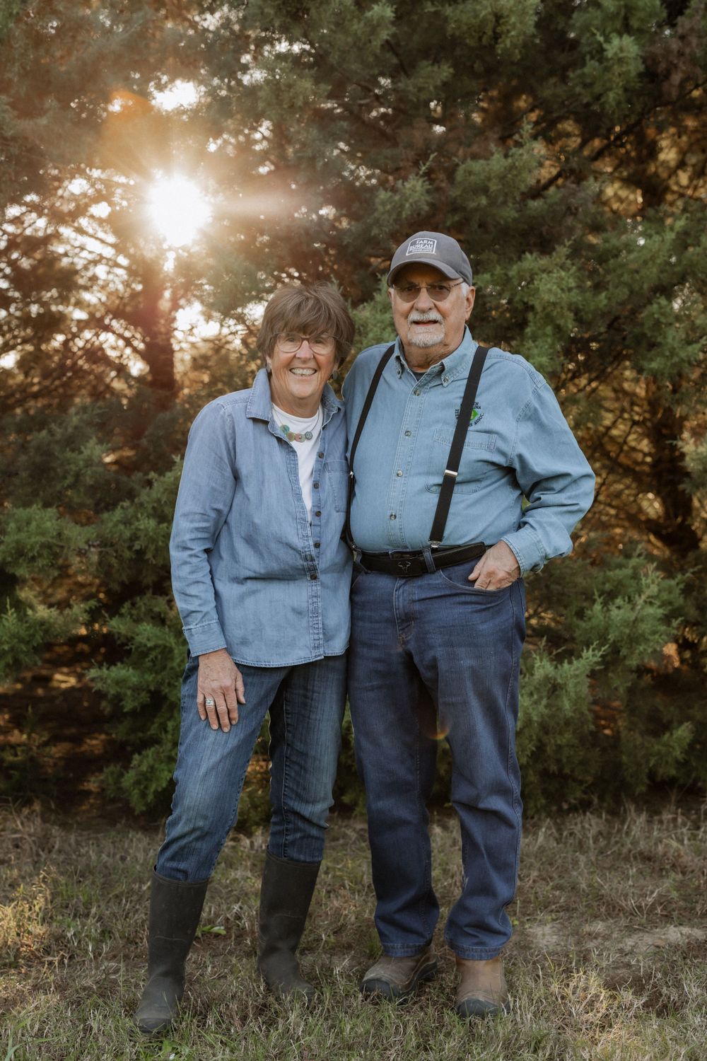 An elderly couple stands together outdoors at sunset with sunlight filtering through pine trees in the background.