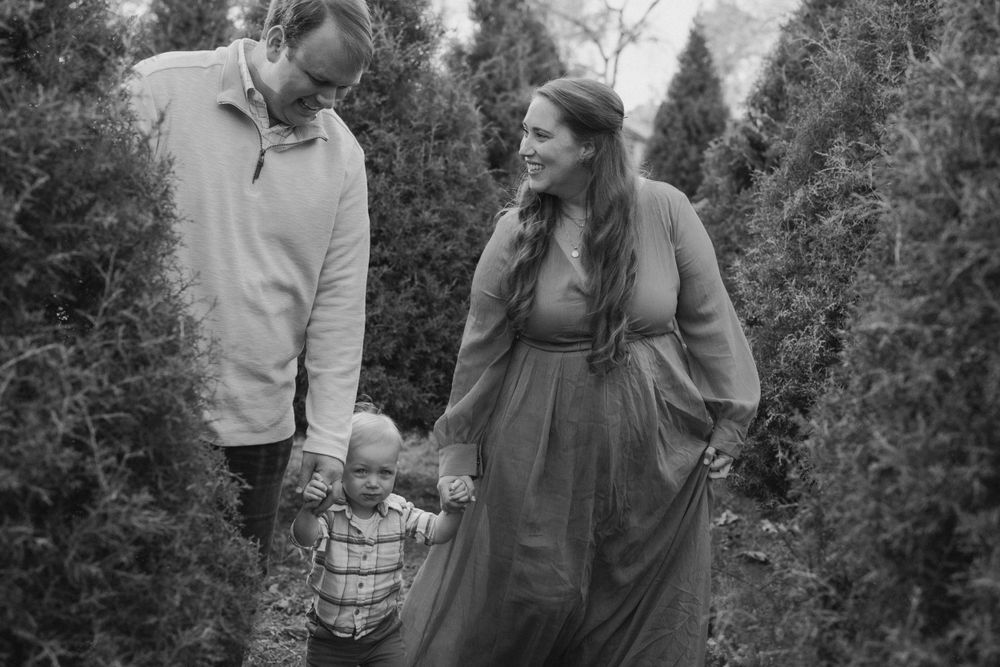 A family in vintage clothing holds hands together while walking through an evergreen tree farm in black and white.