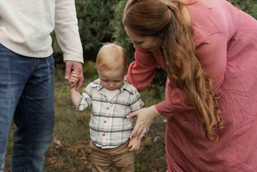A toddler in a striped shirt holds hands with accompanying adults while walking outdoors in a grassy area.