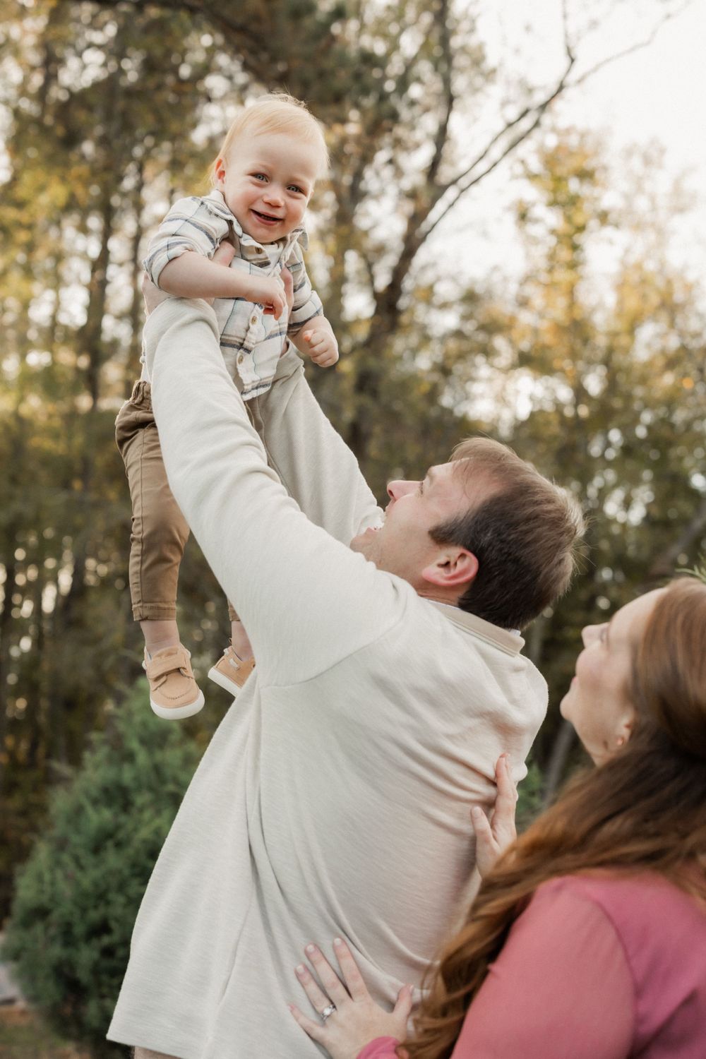 A parent lifts a joyful young child high into the air while standing outdoors with autumn trees in the background.