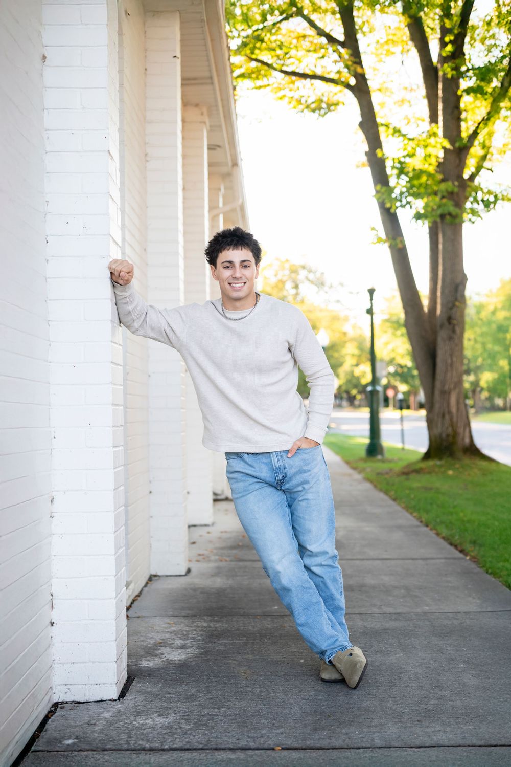 Casual portrait leaning against white column with trees and sidewalk in background.