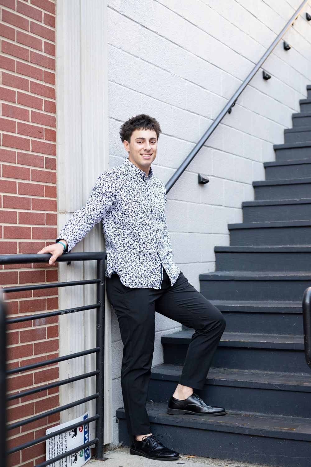 Portrait on dark staircase wearing patterned shirt against white and brick wall.