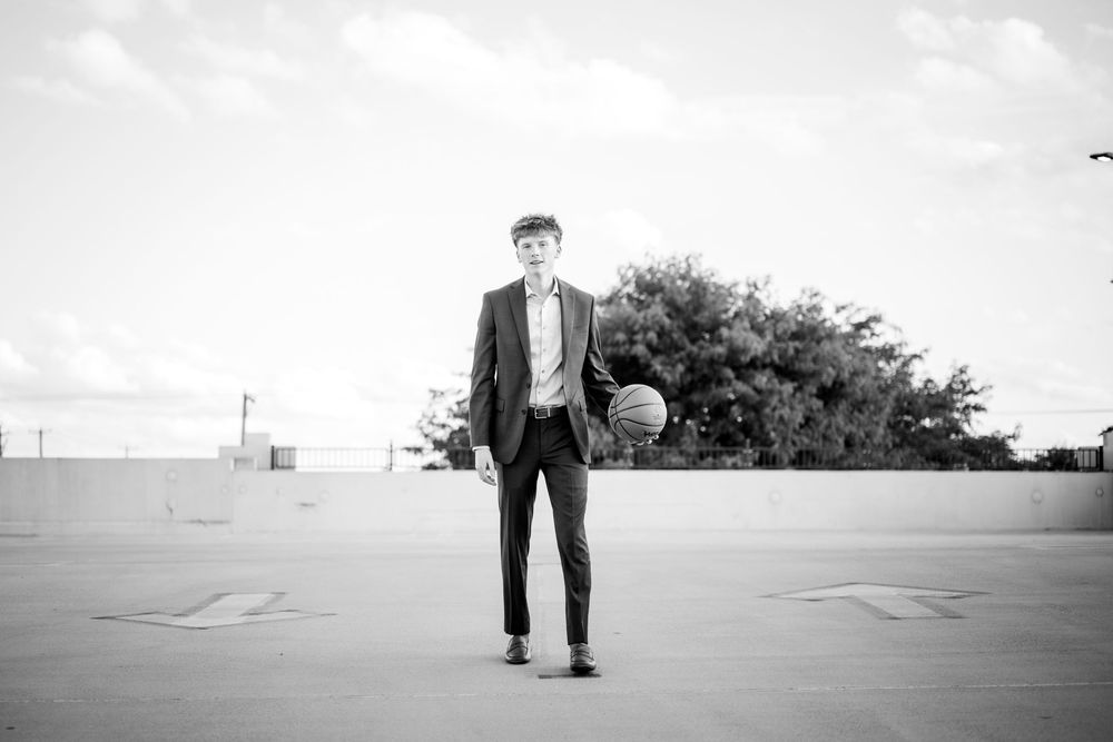 Athletic person in formal attire poses with basketball in parking lot, black and white photo.