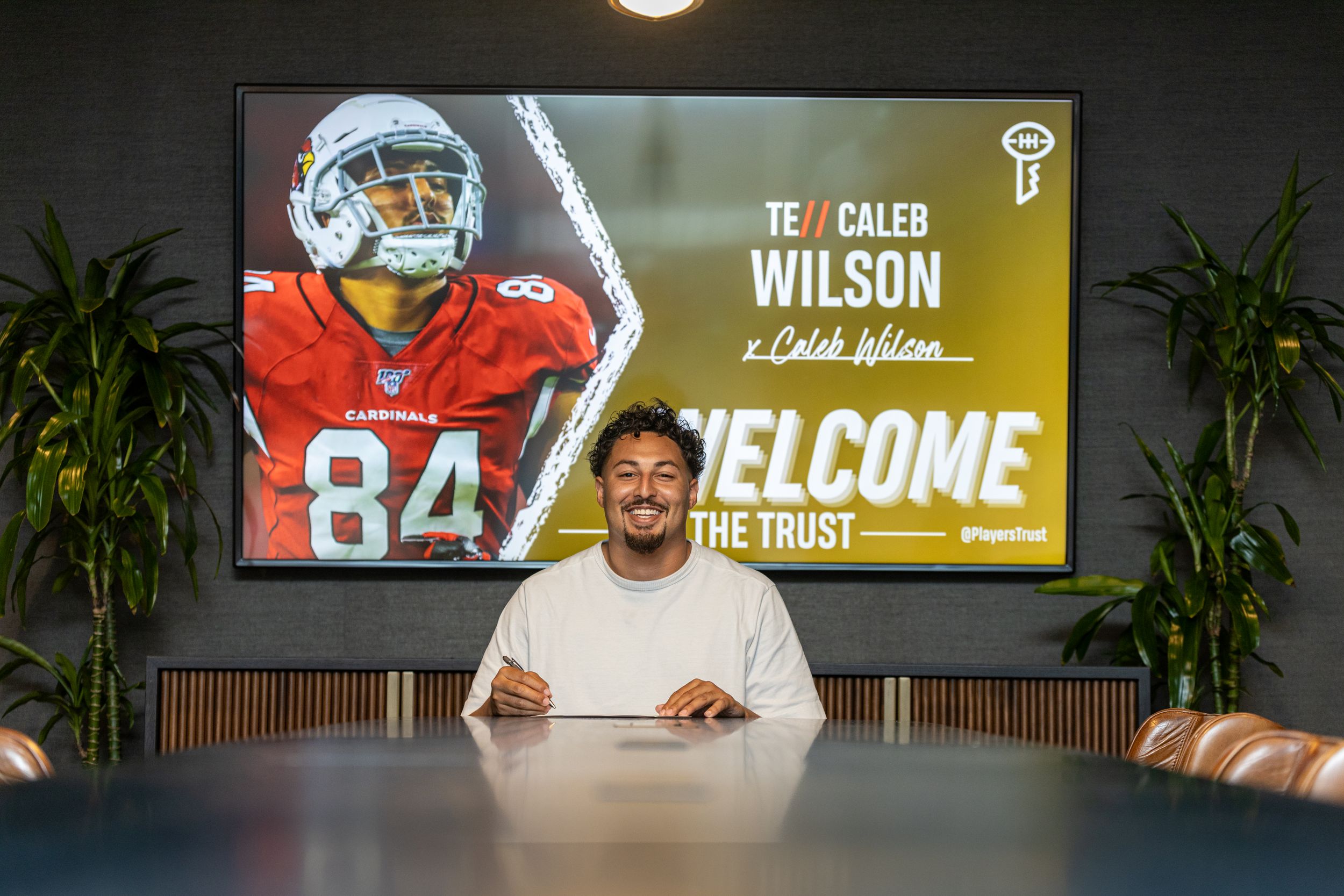 NFL player signs contract at desk with display screen showing jersey number 84 in background.