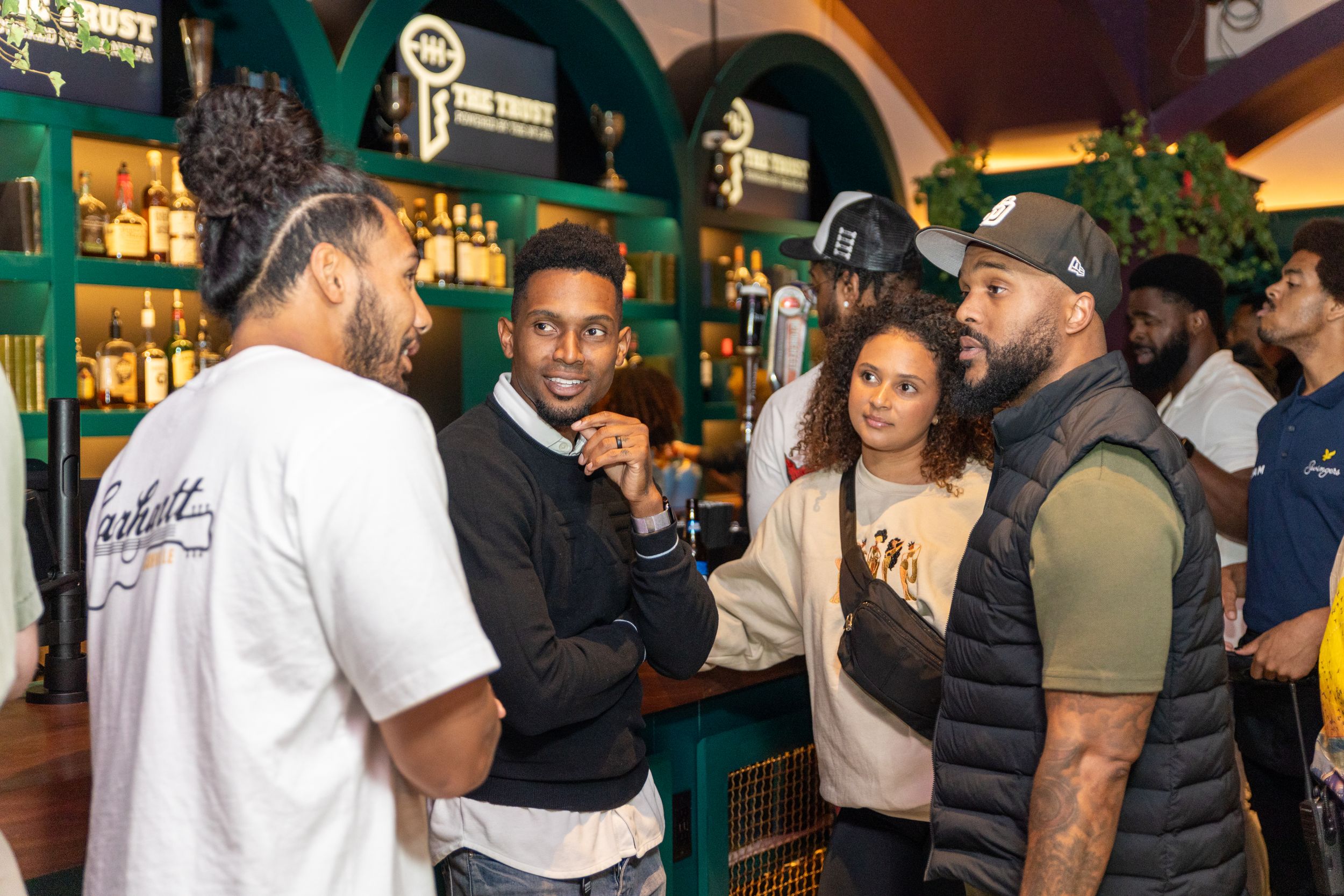Group of people engaged in lively conversation at a bar with green archways.