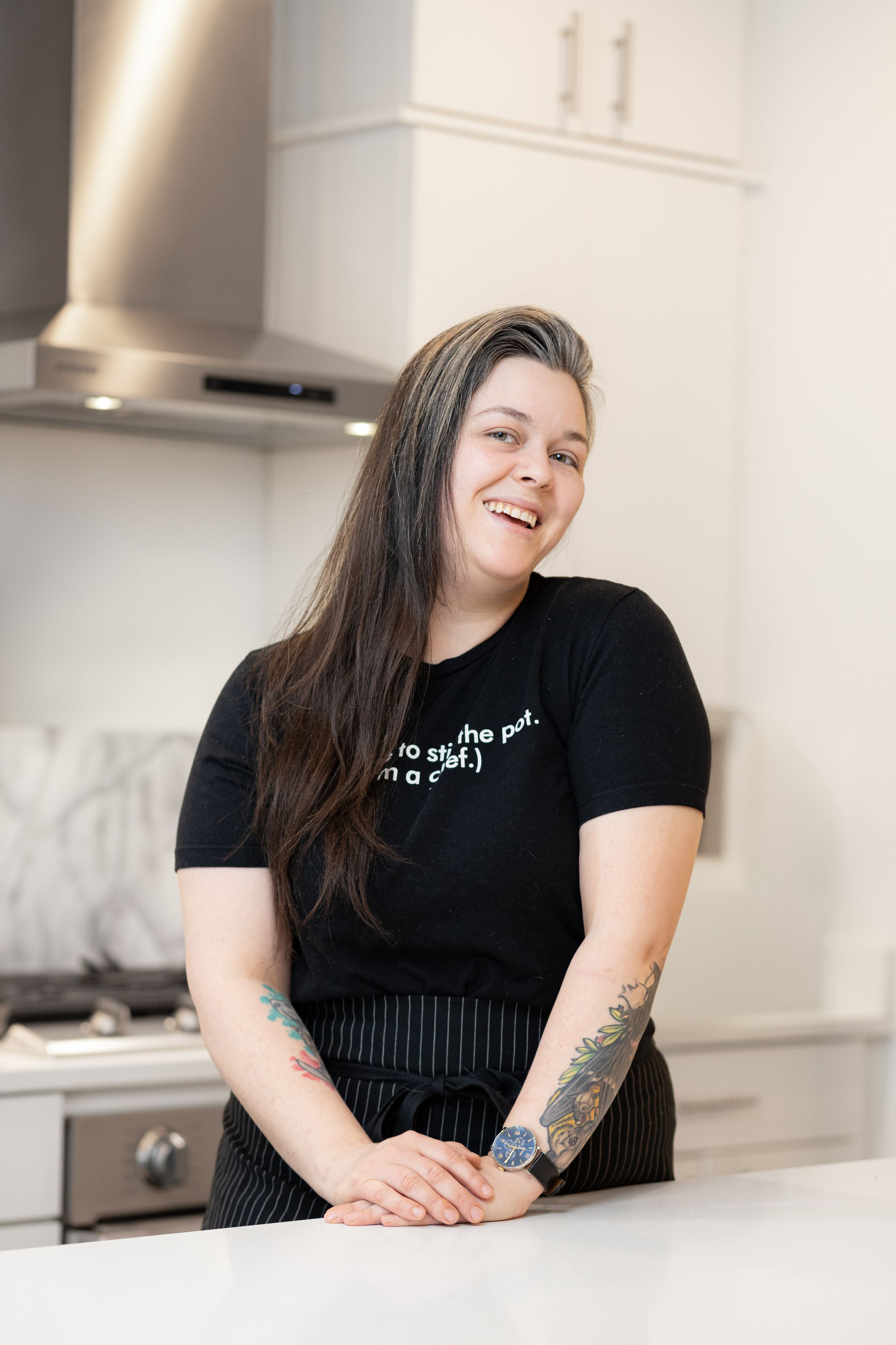 Chef in black attire smiling while standing in a modern white kitchen.