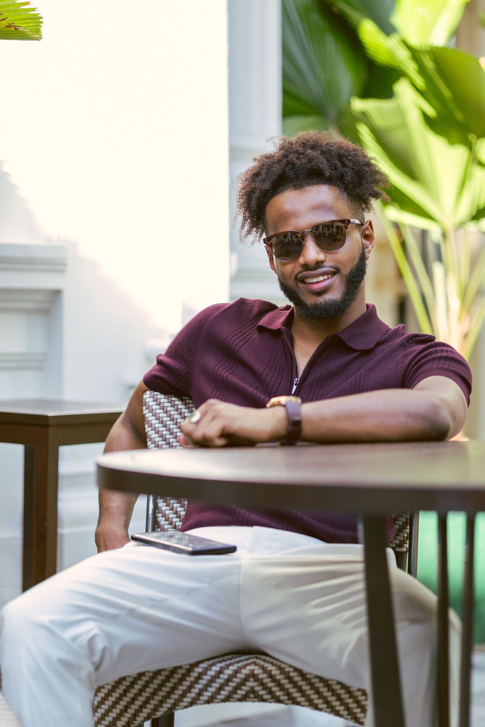 Person relaxing at outdoor cafe table wearing burgundy polo and sunglasses with tropical plants in background.
