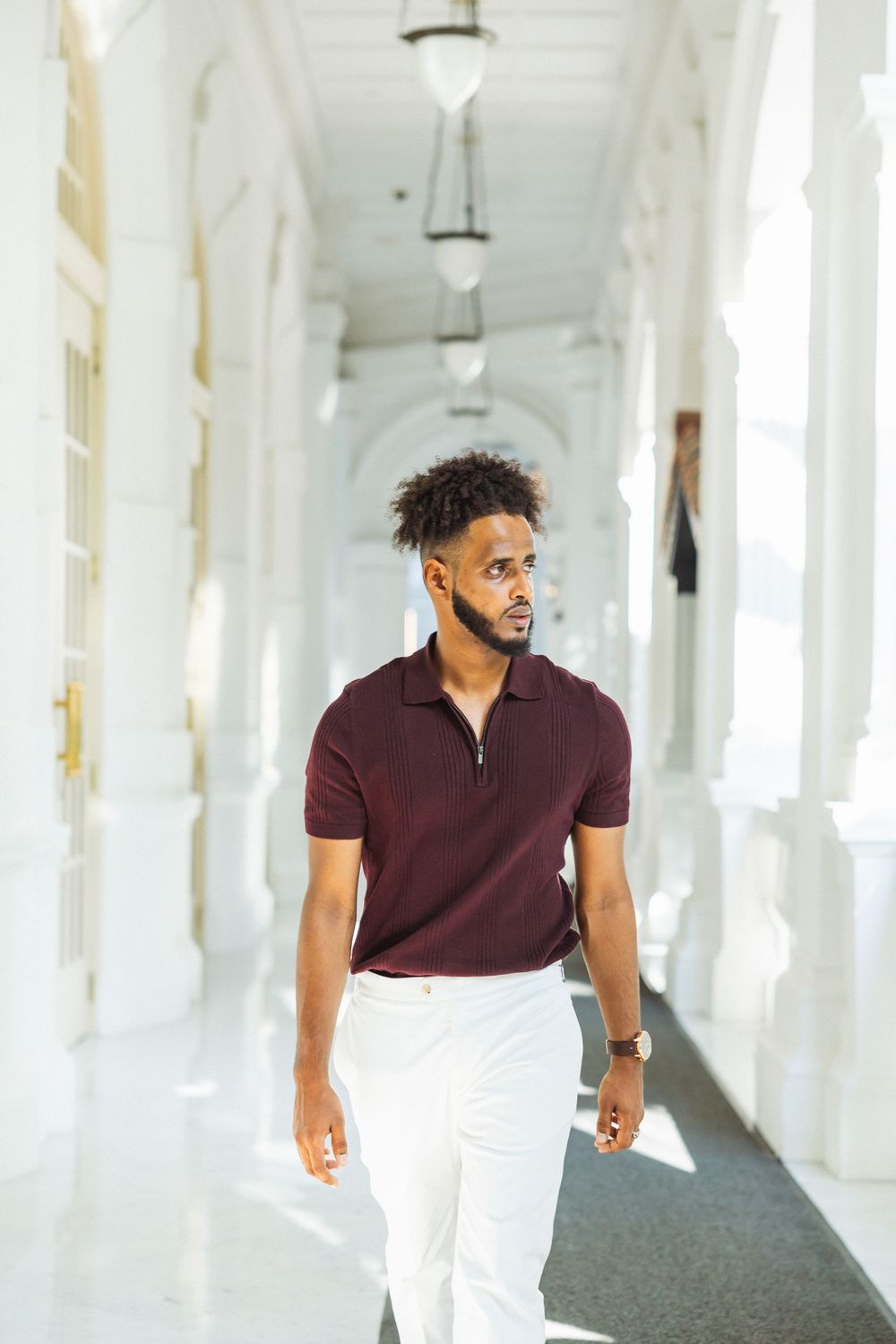 Person walking through bright white hallway wearing burgundy polo shirt and white pants.