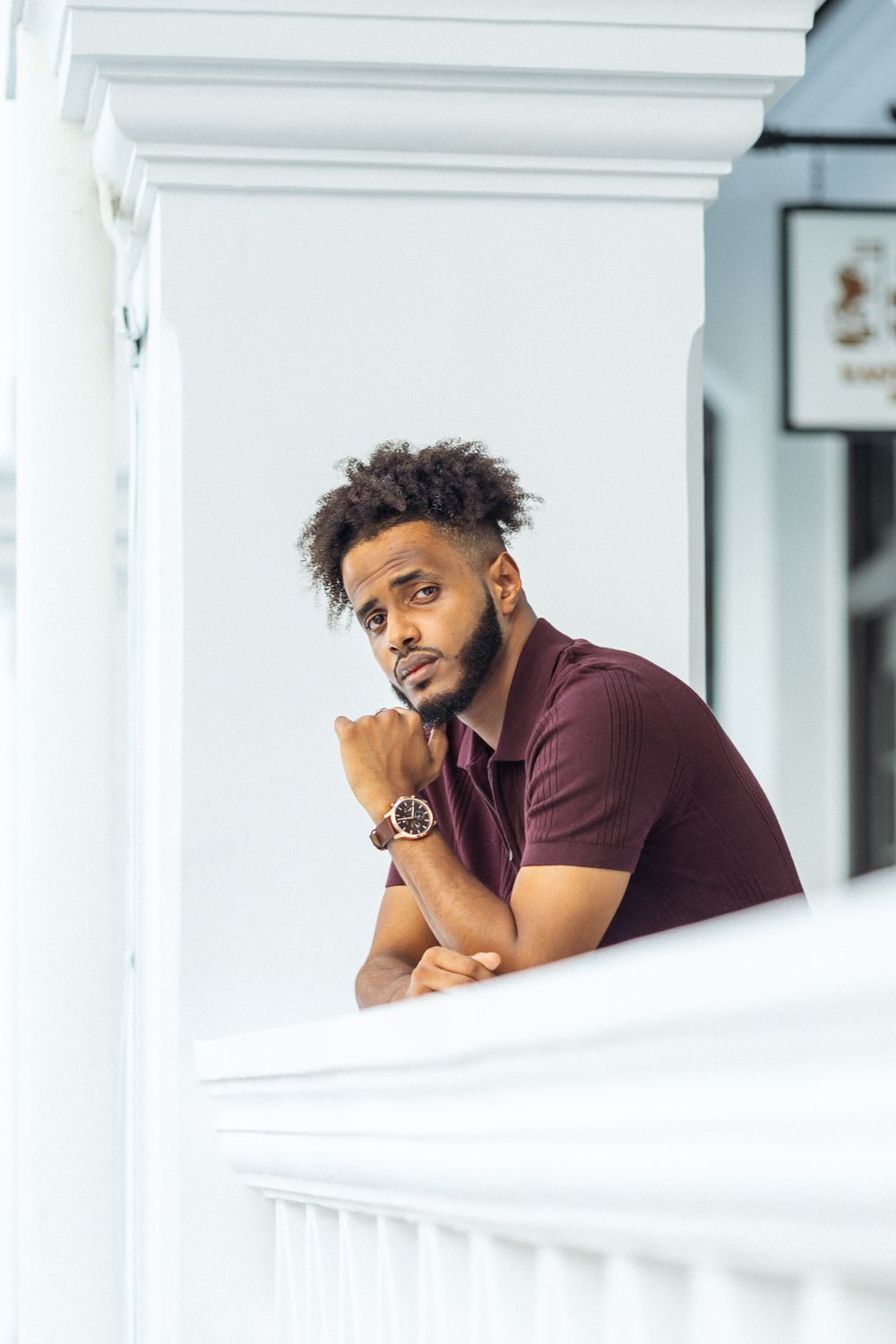 Person in burgundy polo shirt leaning thoughtfully on white railing with classic architecture details.