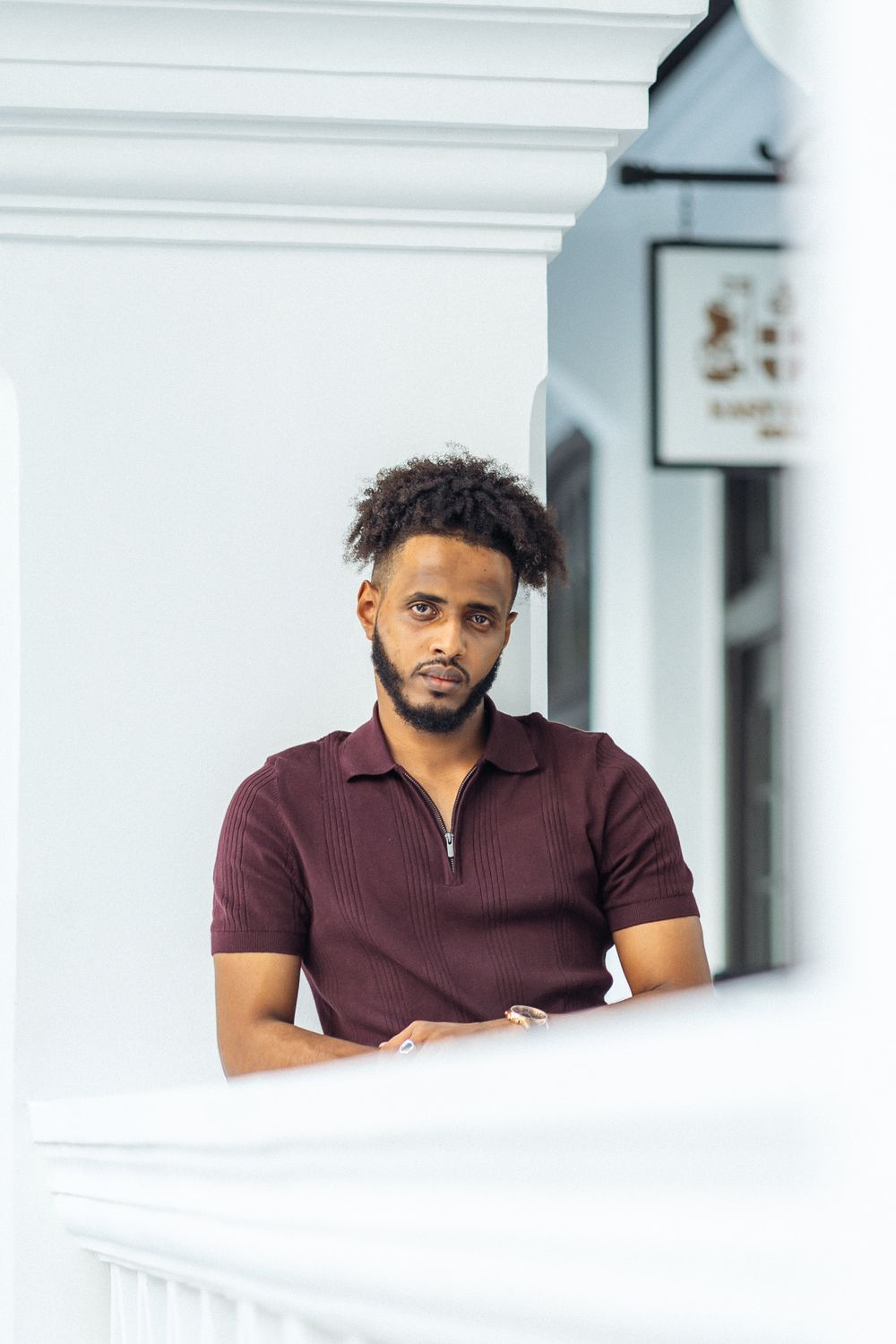Man in burgundy polo leans on white colonial architecture balustrade while looking at camera.
