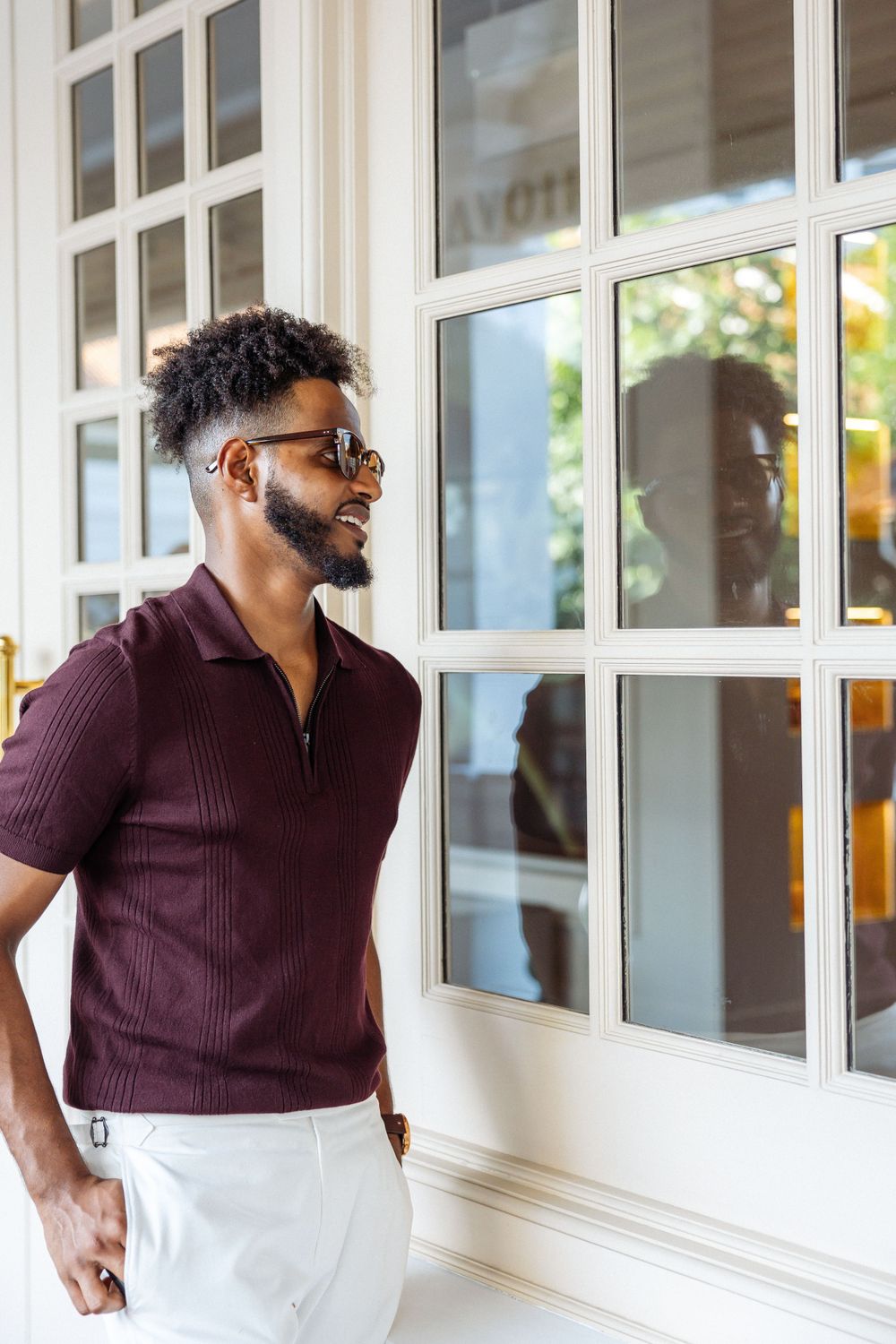 Stylish man in burgundy polo and white pants looks through window while smiling.