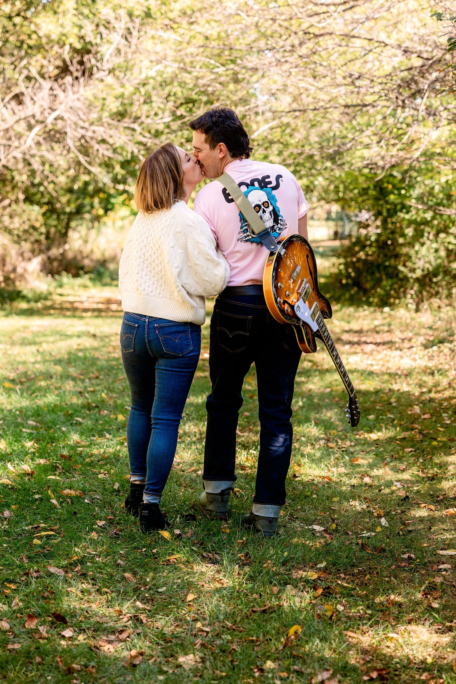 Couple kisses while walking through a Novi park, the husband has a guitar slung over his back. Rachel Martinez Photography