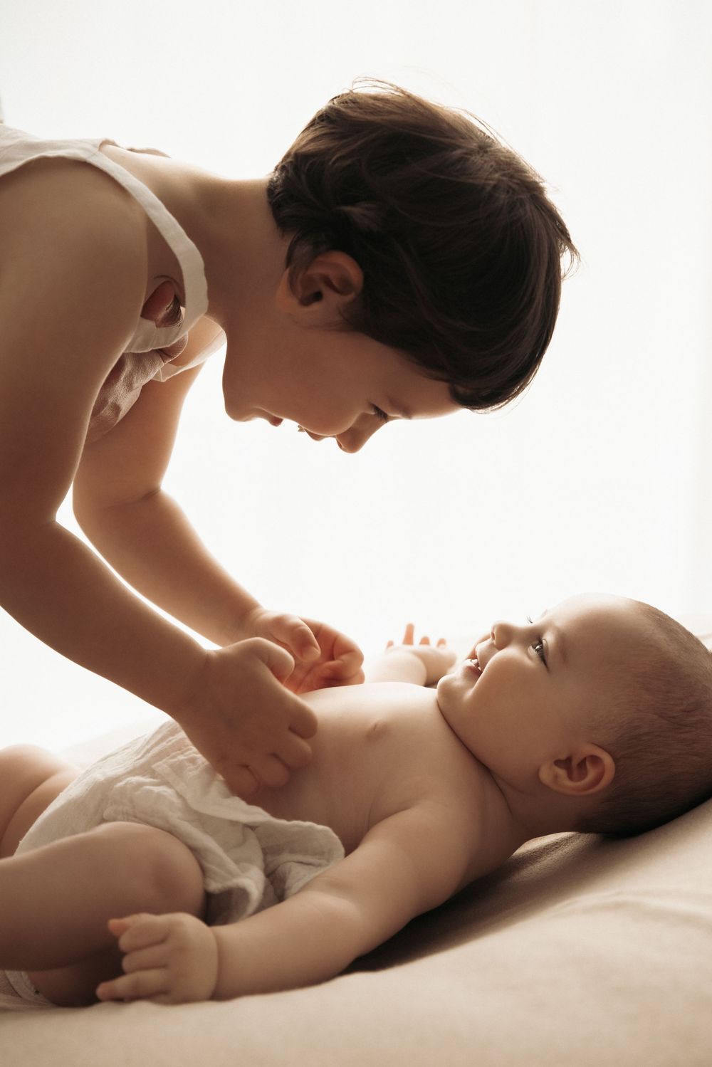 A tender moment between parent and baby while snuggling and playing together on white bedding in soft lighting.