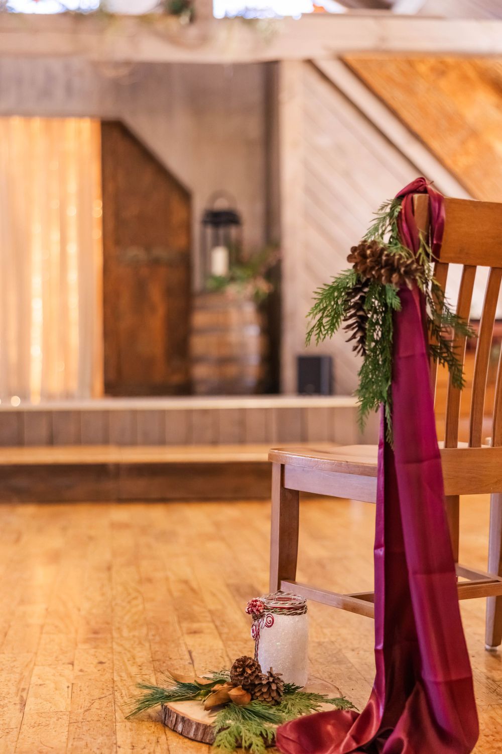 A wooden chair decorated with burgundy fabric and evergreen branches in a rustic indoor setting.