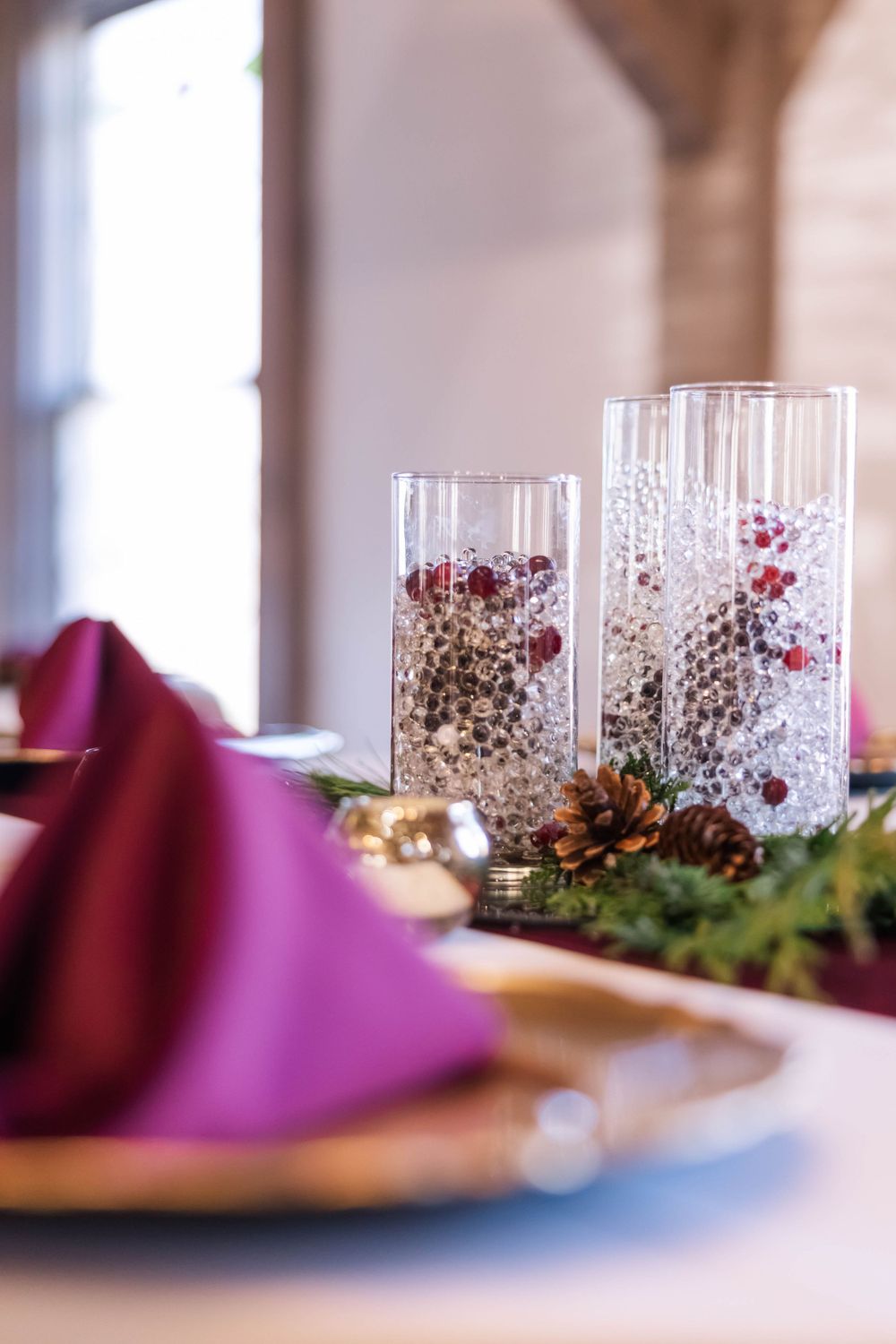 Elegant table setting with burgundy napkins and glass cylinders filled with silvery winter decorations.