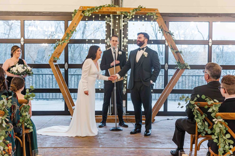 Wedding ceremony takes place under a wooden hexagonal arch decorated with greenery against floor-to-ceiling windows.