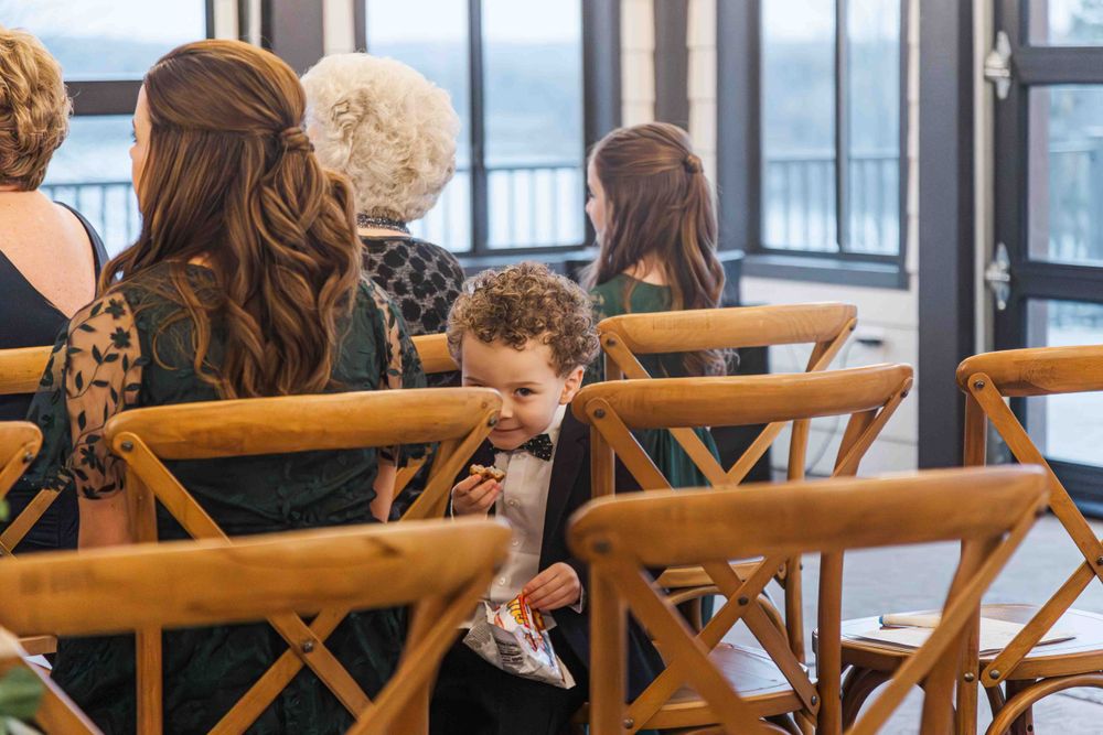 A young child peeks between wooden cross-back chairs during an indoor gathering or event.