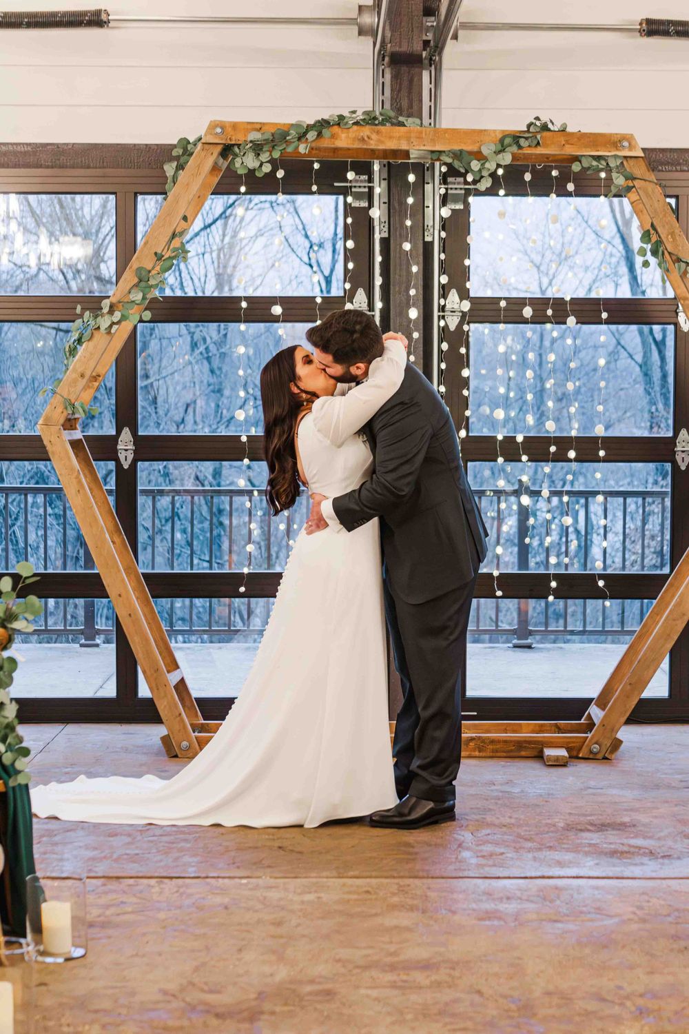 A couple embraces during their winter wedding ceremony under a wooden hexagonal arch decorated with greenery and lights.