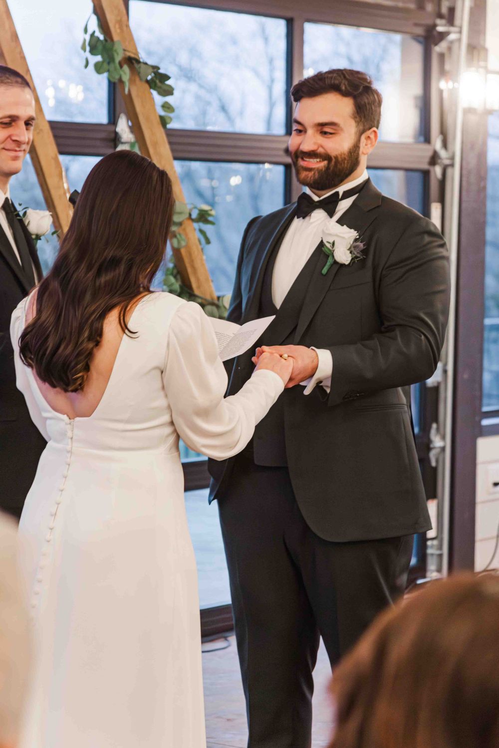 A groom in a black tuxedo and bride in a white dress hold hands during their wedding ceremony near large windows.