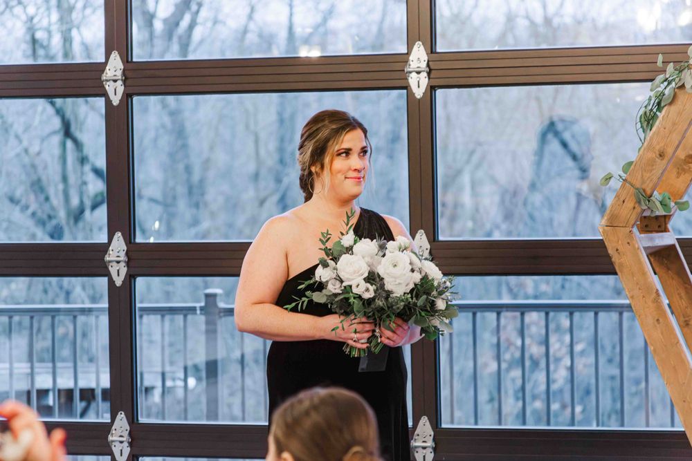 A member of the wedding party stands at the front holding a white floral bouquet in front of a glass-walled venue.