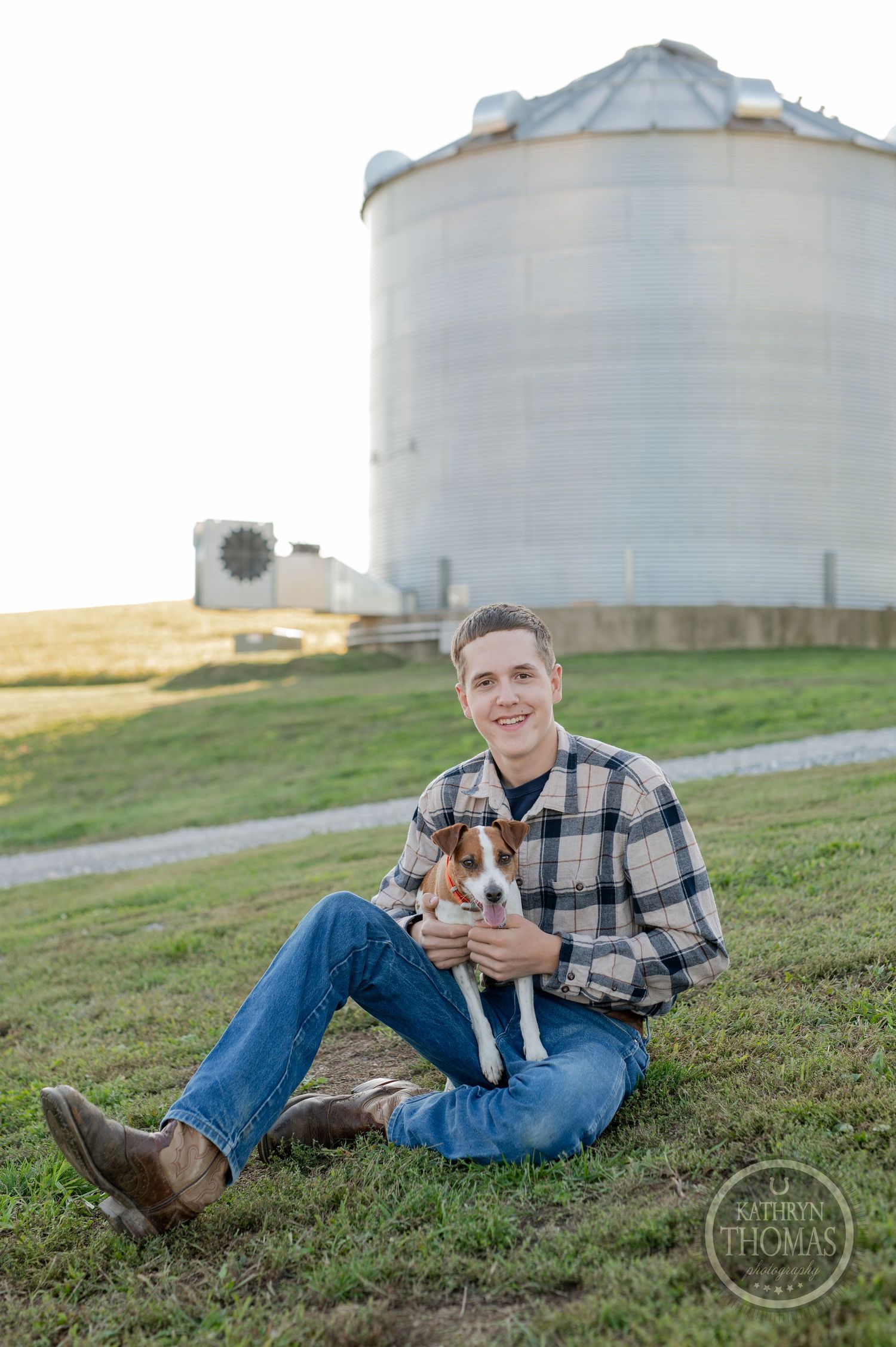 Person in plaid shirt sits with small dog in front of grain silo at sunset.