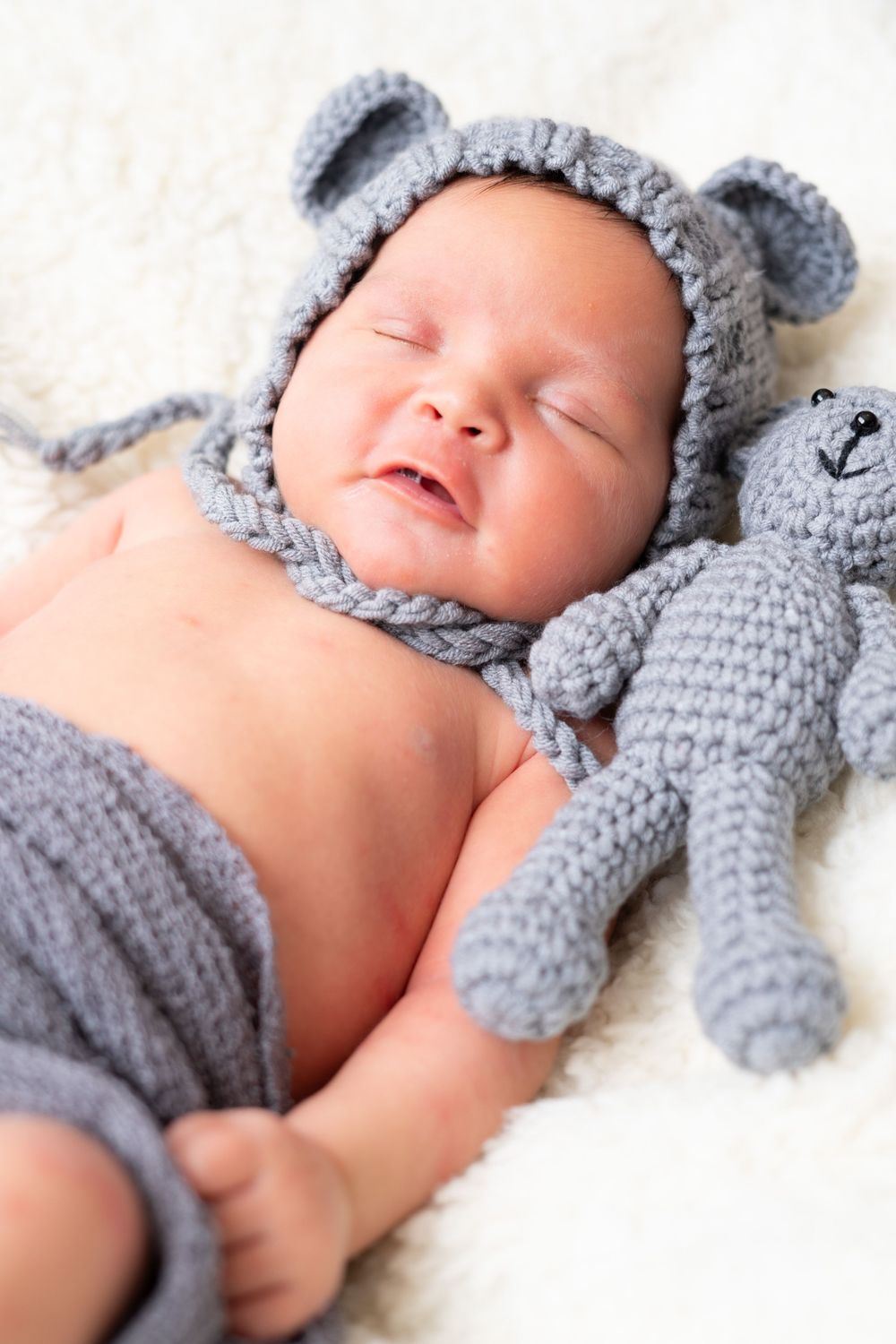 Sleeping baby in gray knit bear hat lying on white blanket with matching gray teddy bear toy.
