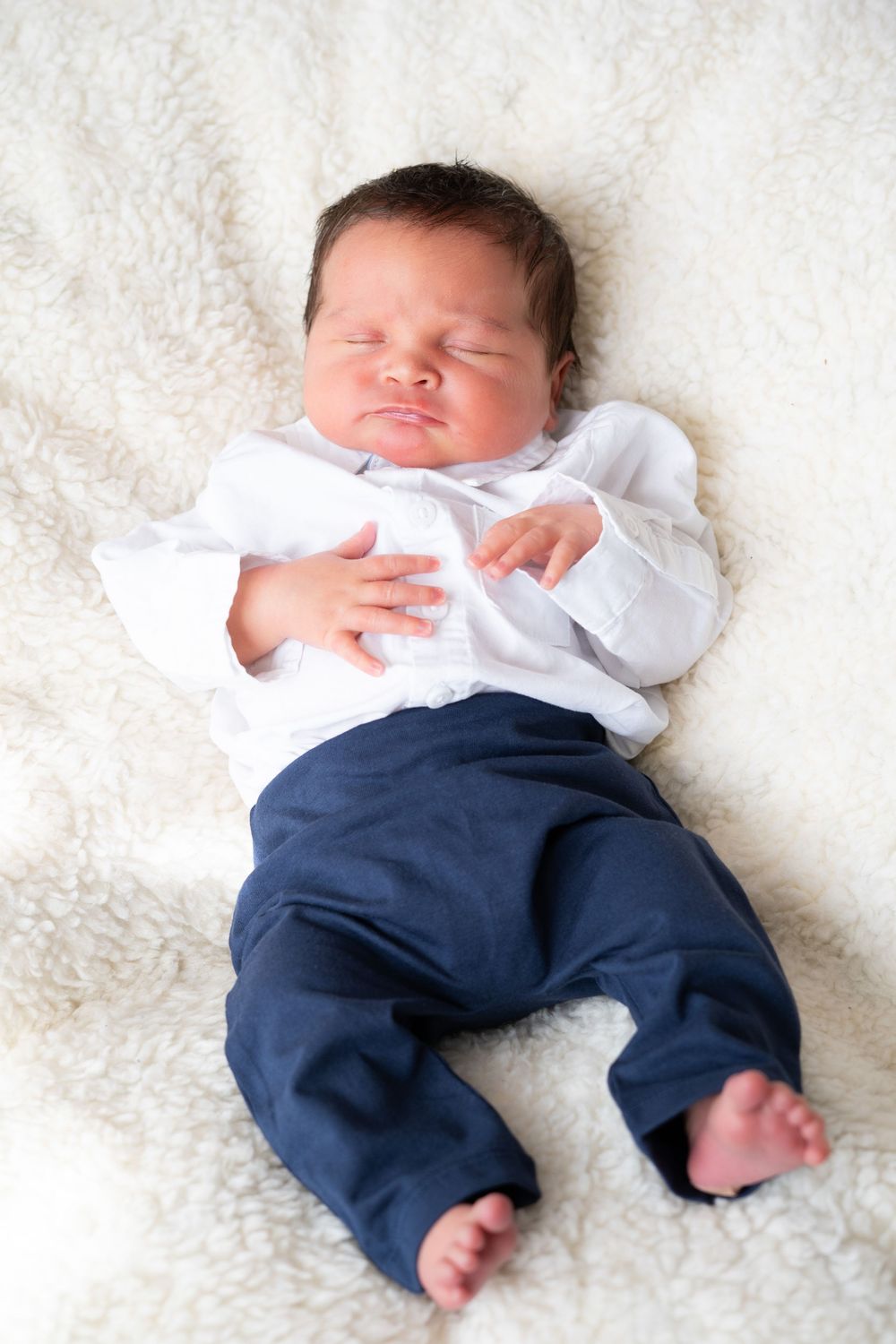 Sleeping newborn baby wearing white shirt and navy pants lies on soft white blanket.
