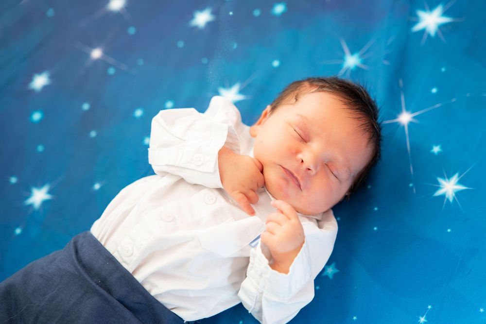 Sleeping newborn in white top and navy pants lies peacefully against twinkling blue starry backdrop.