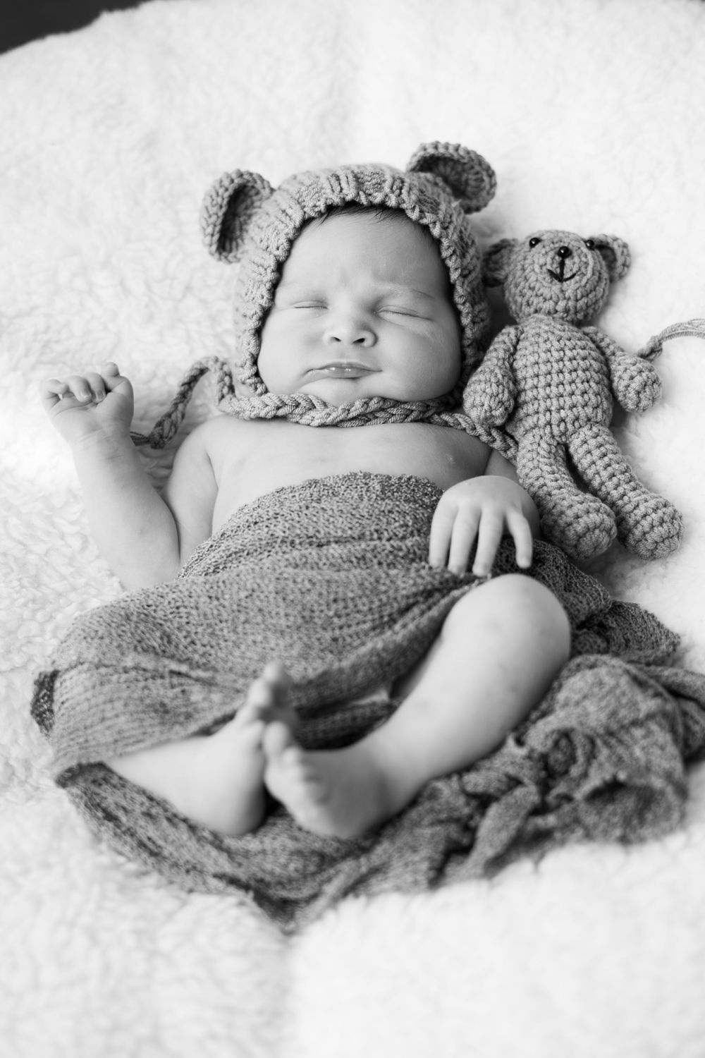 Black and white photo of sleeping baby wearing knit bear hat lying on blanket with matching teddy bear toy.