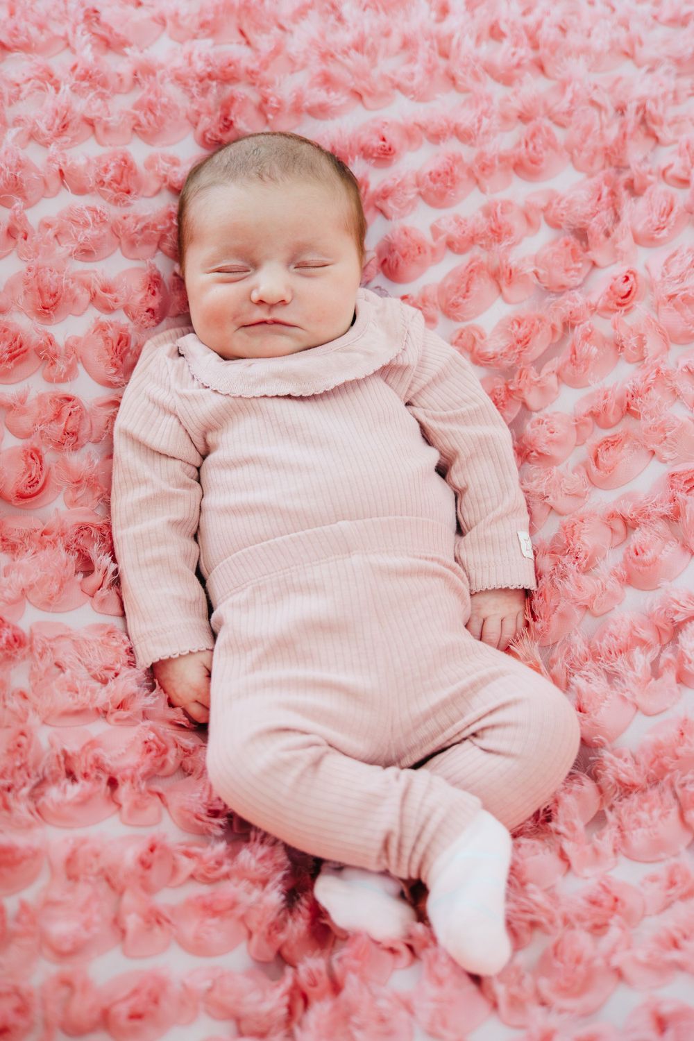 Sleeping baby wearing pink ribbed outfit lying on soft pink textured blanket during newborn photoshoot.