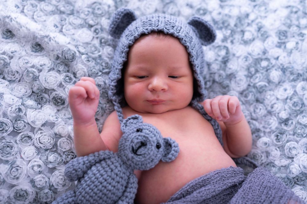 Sleeping baby wearing knit bear hat with matching stuffed toy on patterned blanket.