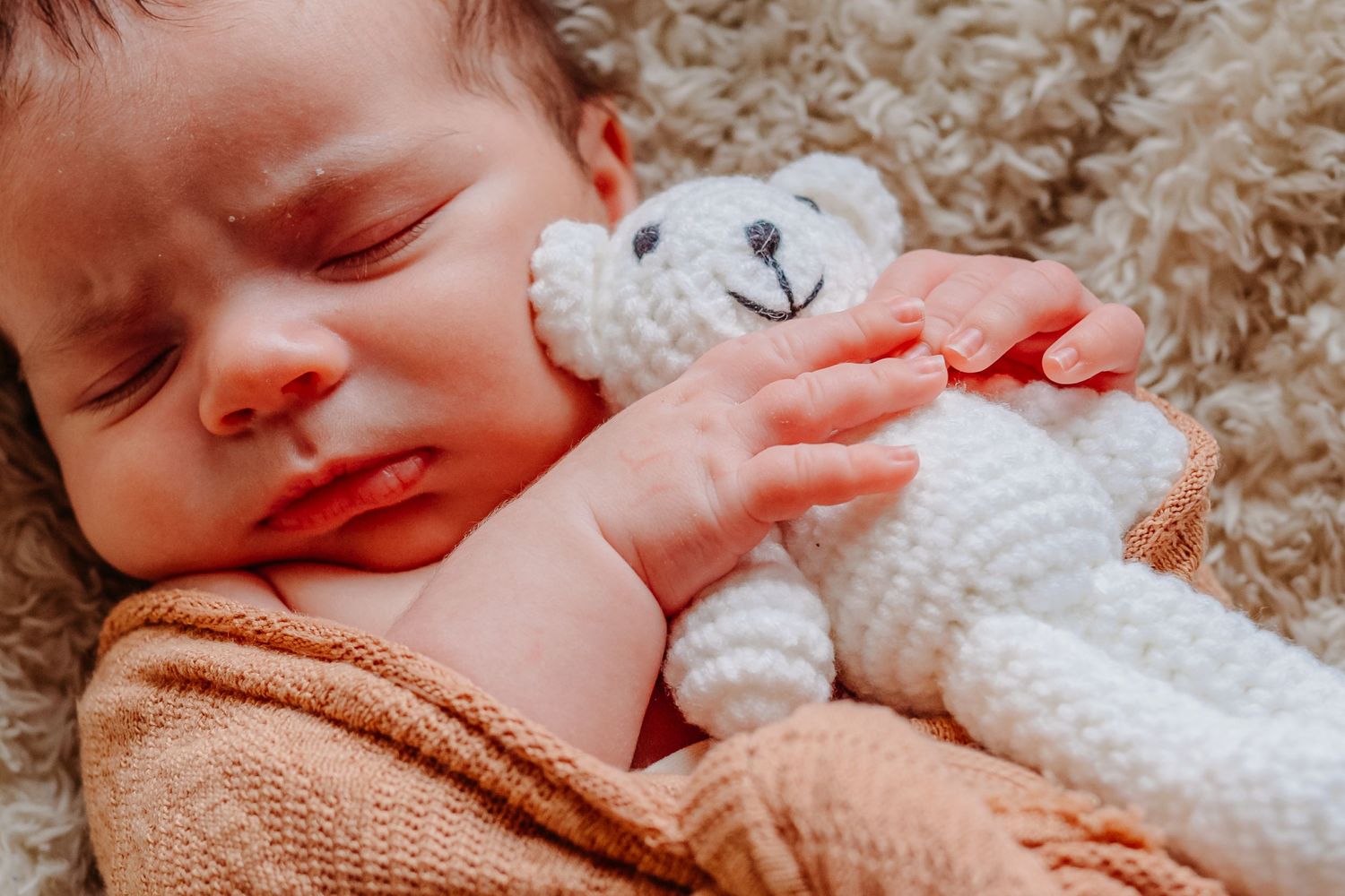 Close-up of baby sleeping peacefully with white knitted teddy bear toy on beige blanket.
