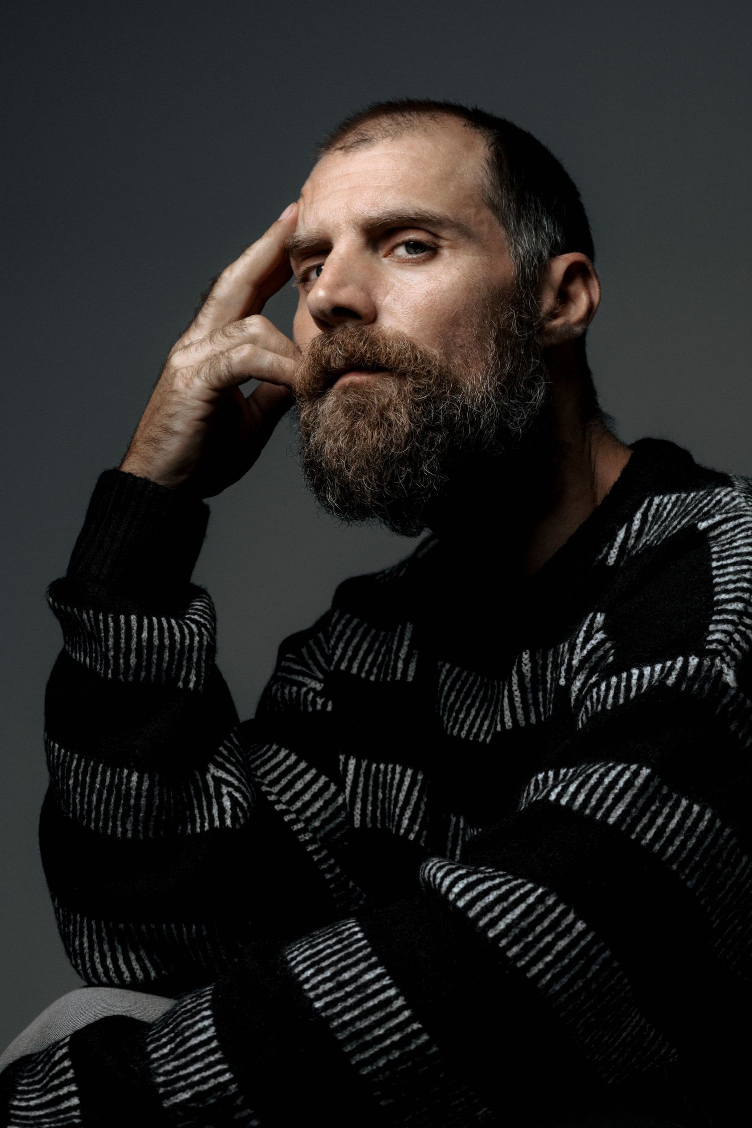 Dramatic studio portrait of Jose Manuel Poga in black and white striped sweater with hand near temple against dark background.