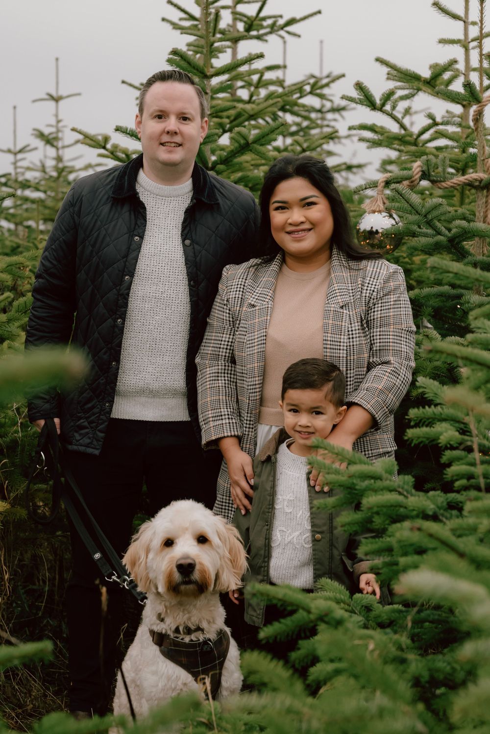 A family poses with their fluffy white dog among Christmas trees at a tree farm during the holiday season.