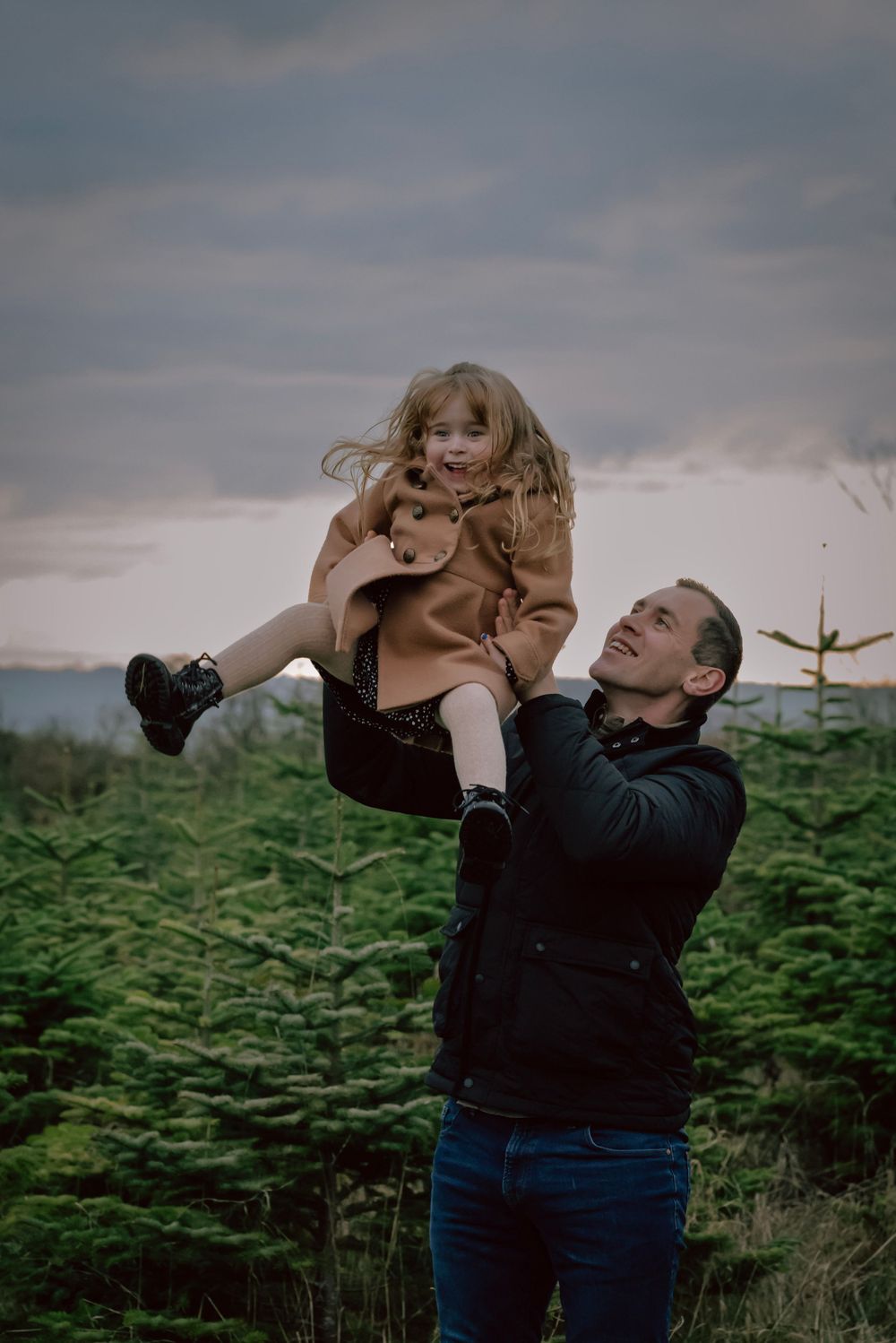 A father and his daughter play in the christmas trees. He throws her to the sky as she giggles and smiles.