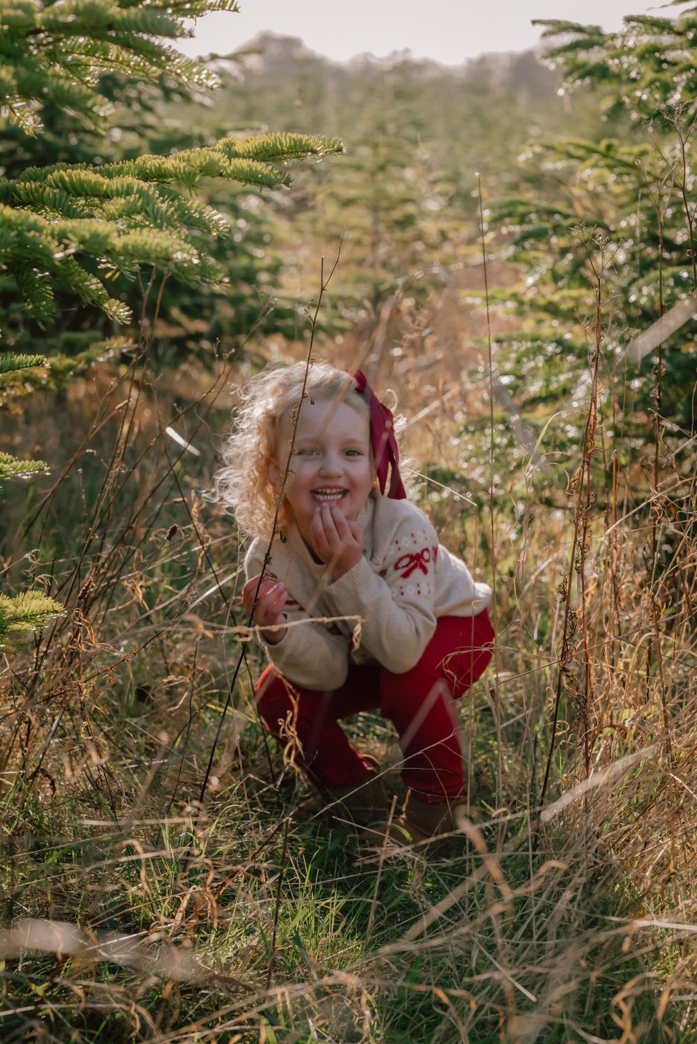 A young girl with blonde curly hair is caught in her hiding place amongst the trees. She giggles as she crouches in the long grass.