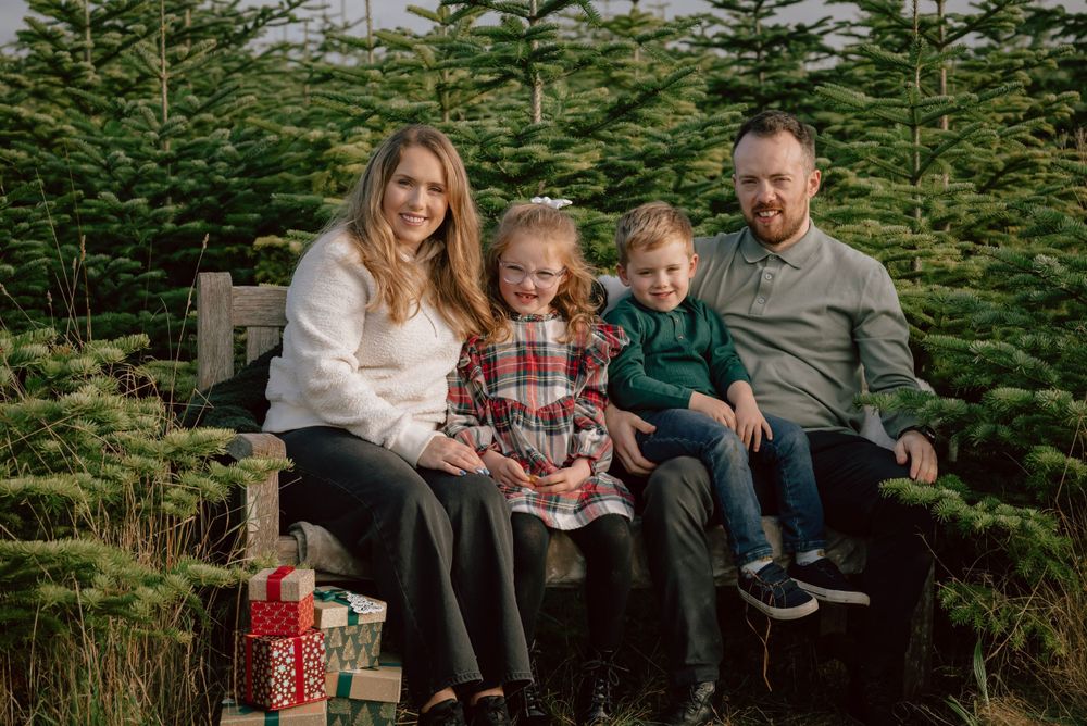 A mother, son, daughter and father sit on a wooden bench in the winter sunshine, surrounded by christmas trees.