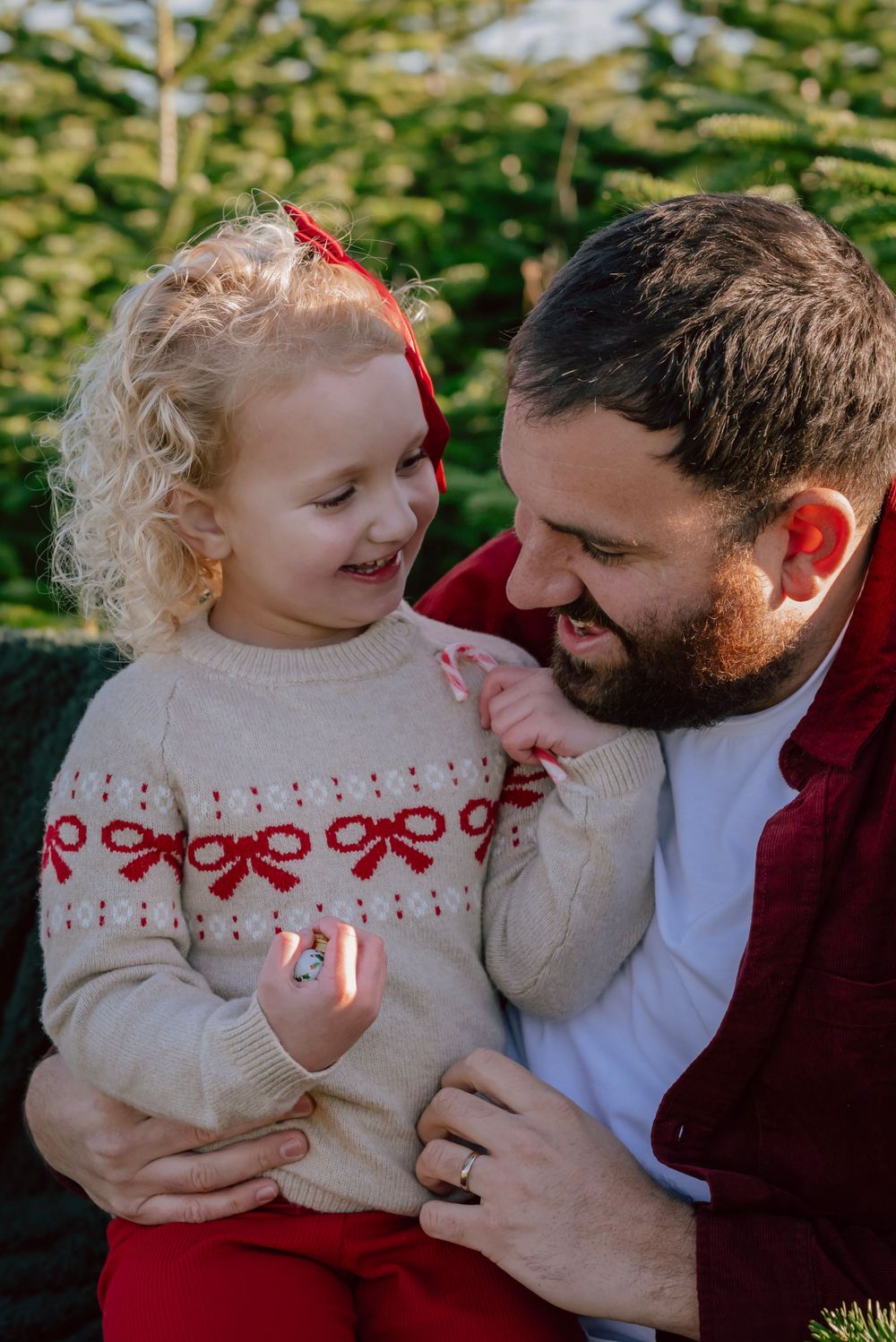 A girl sites with her father wearing a beige jumper adorned with a red bow pattern. The sun is streaming through the trees.