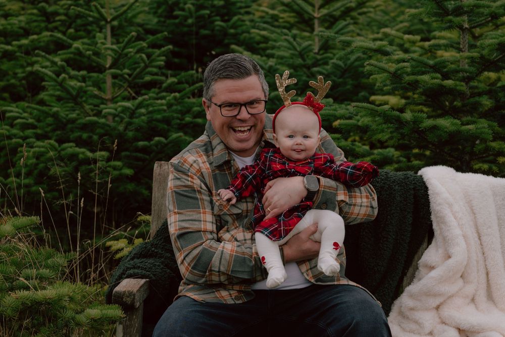 A festive holiday scene at a Christmas tree farm with cozy plaid attire and evergreen trees in the background.