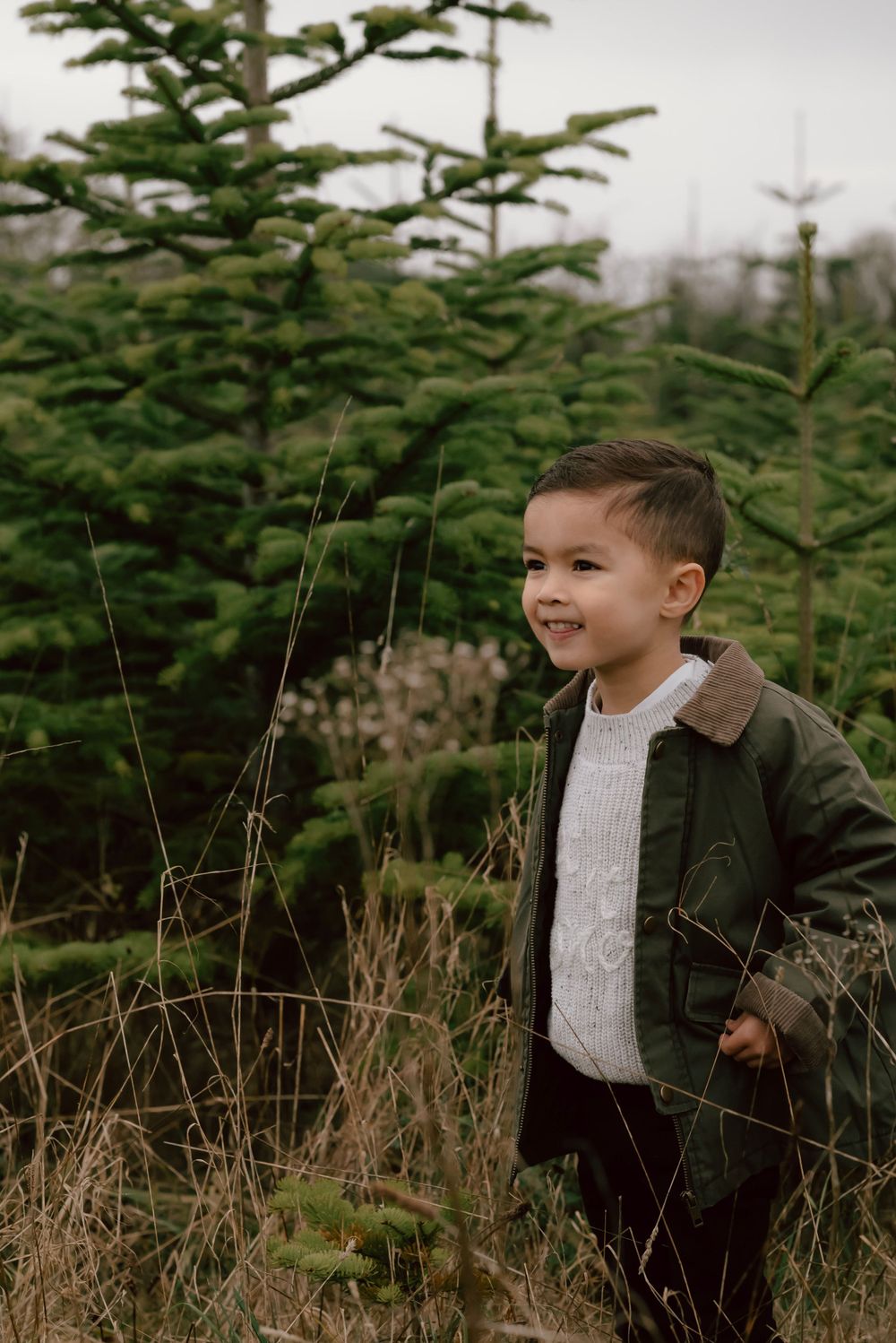 A young child in a green jacket and sweater stands outdoors near evergreen trees on a cloudy day.
